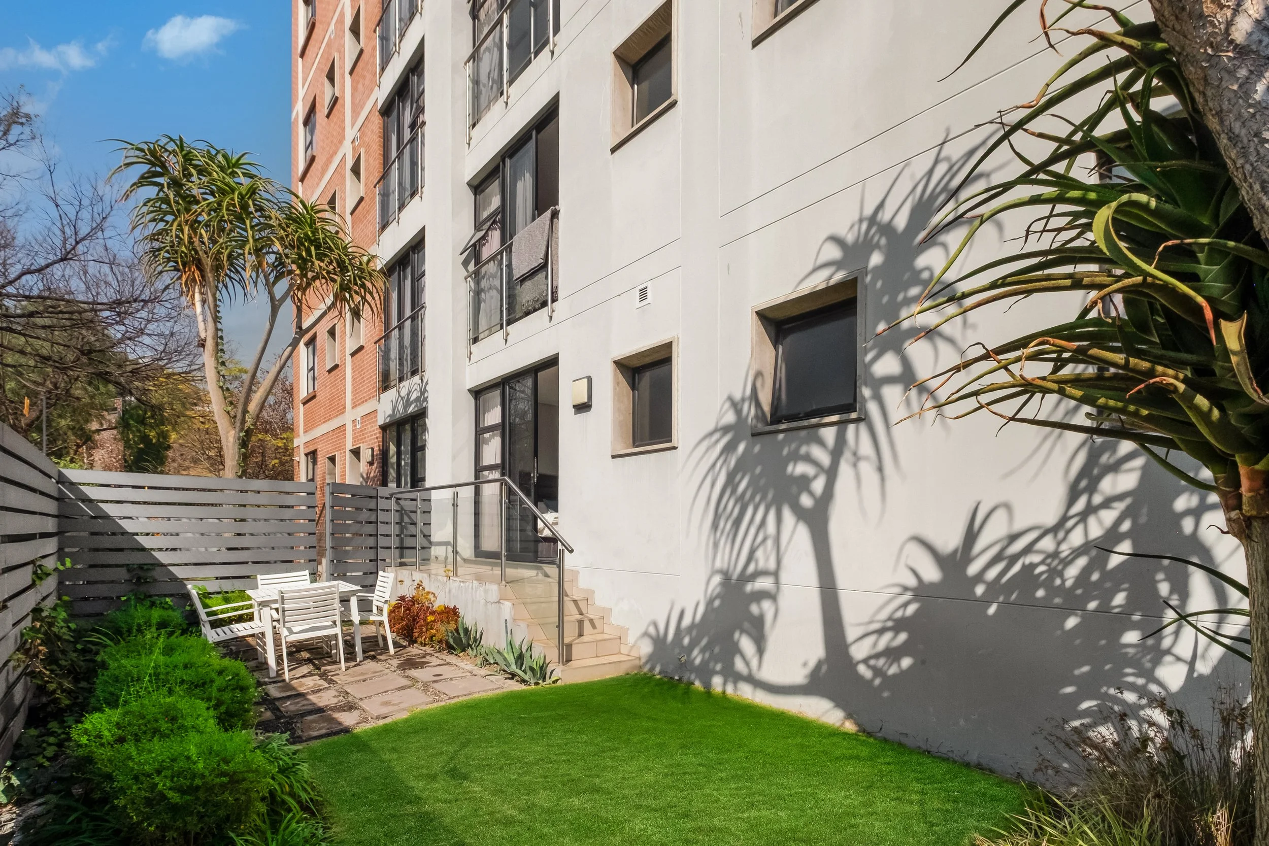 A modern apartment building with several windows, surrounded by a landscaped garden with trees, bushes, a grassy lawn, and a patio area with white chairs and a small staircase.