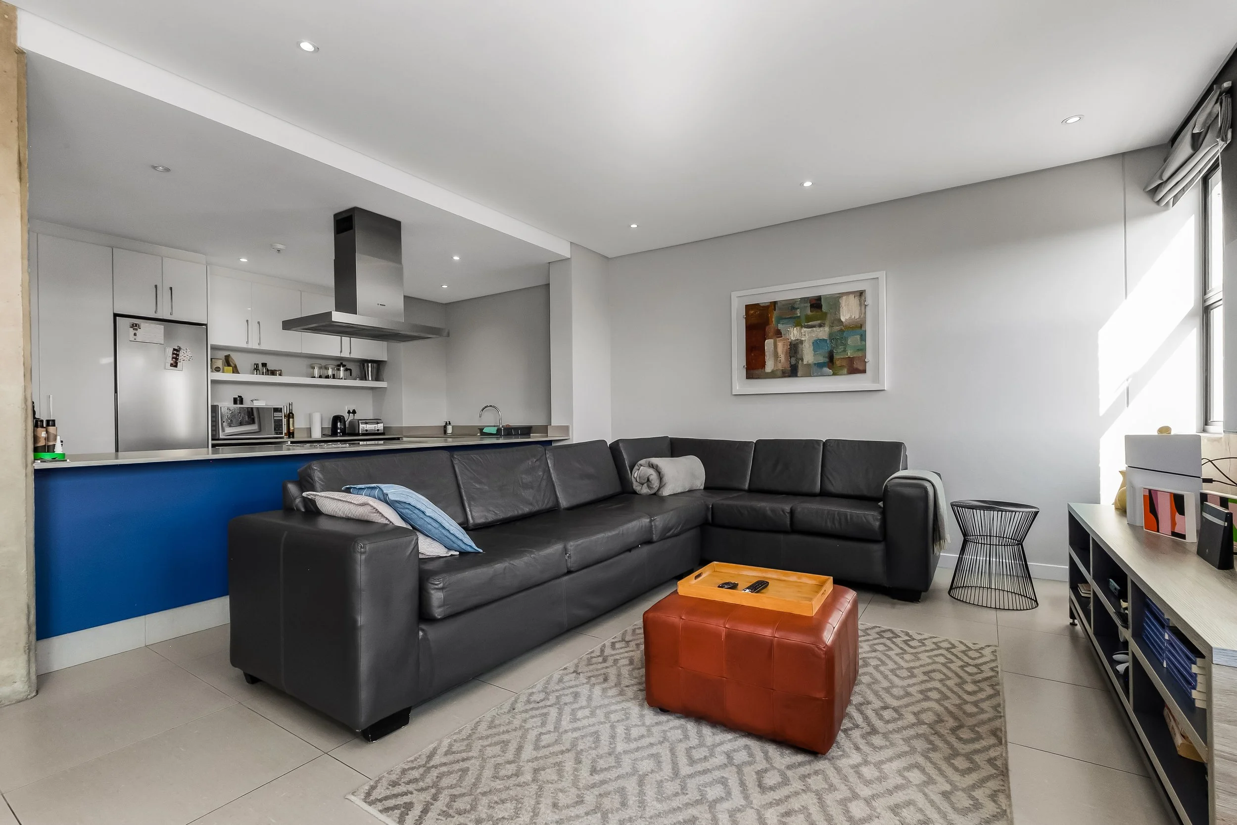 Living room with black leather sectional sofa, orange leather ottoman, white walls, abstract art, and open kitchen with white cabinets and stainless steel appliances.