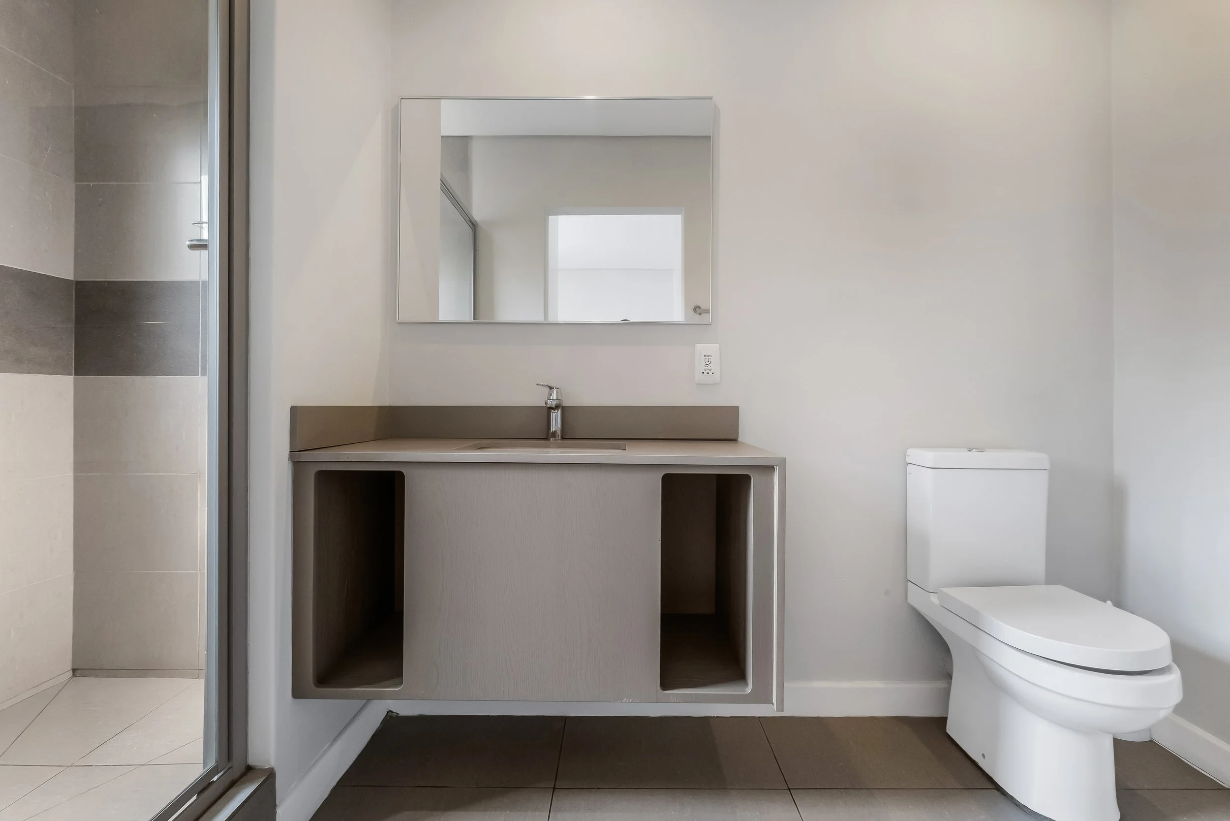 Minimalist bathroom with a white toilet, gray vanity with an open shelf, mirror, and a shower with sliding door on the left.