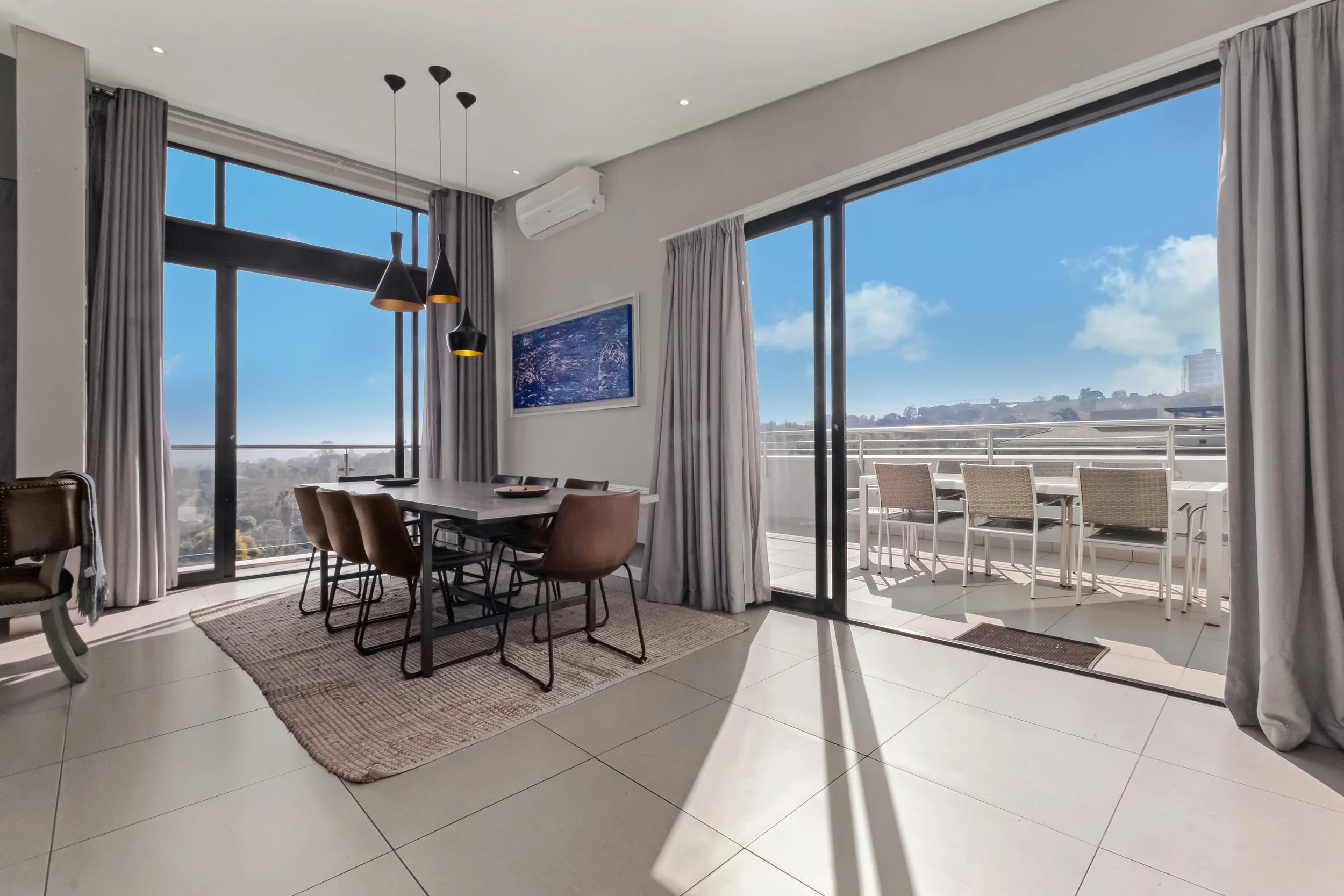 Modern living room with large windows and sliding glass doors, beige tiled floor, dining table with chairs, hanging pendant lights, framed artwork on the wall, and balcony with outdoor seating, overlooking a blue sky and distant buildings.