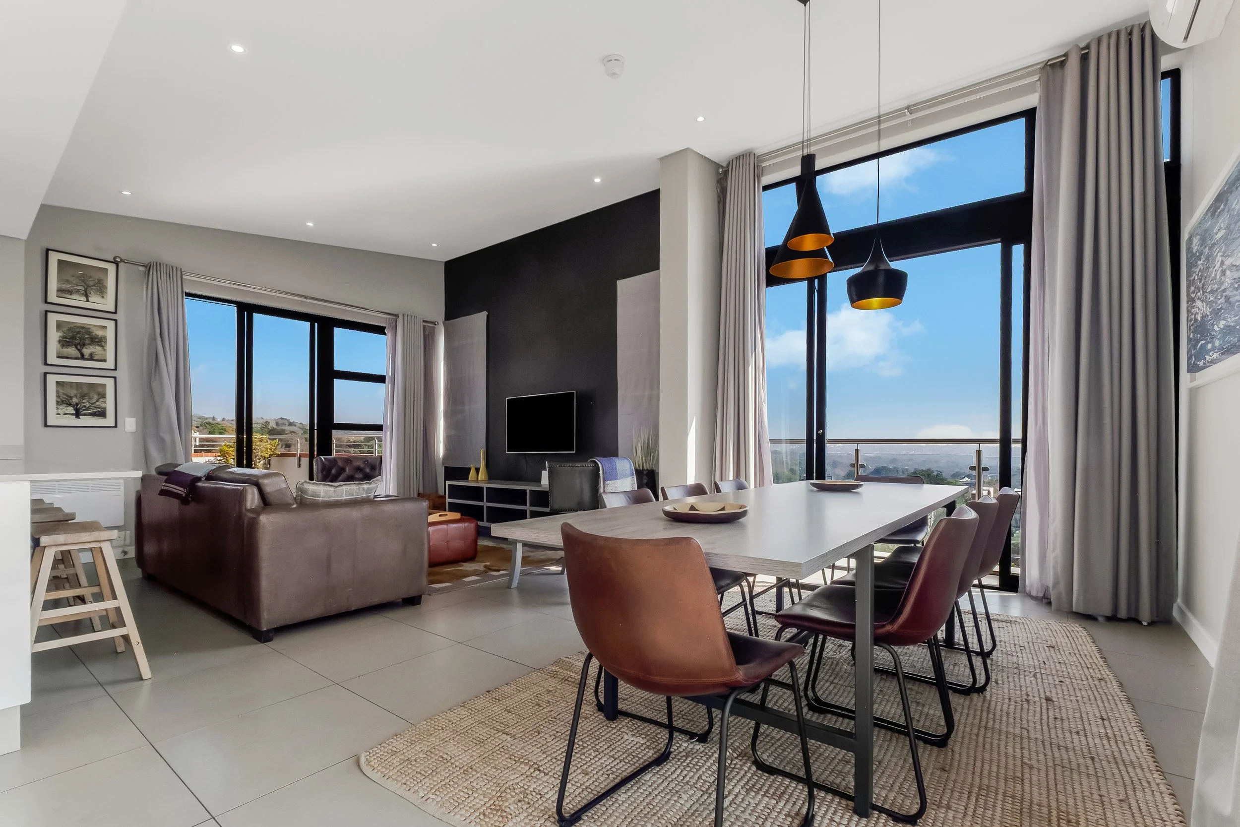Modern living room with large windows showing a blue sky, with a dining table and various chairs, a sofa, wall art, and curtains.