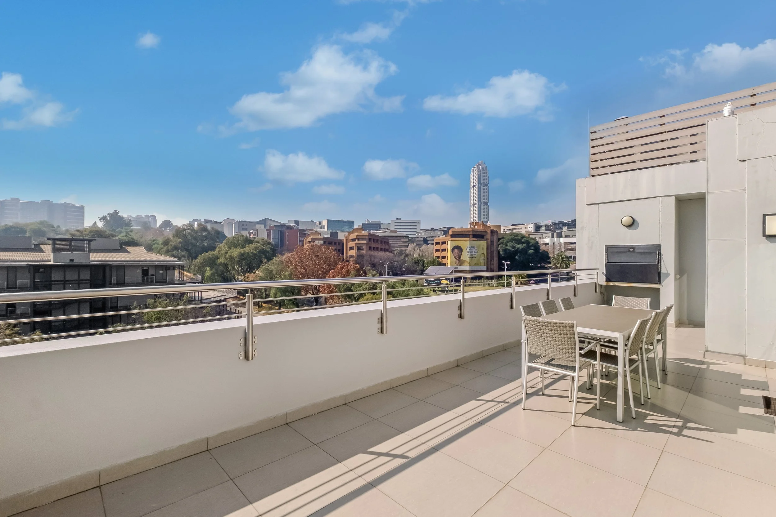 View from a balcony overlooking an urban cityscape with buildings, trees, and a blue sky with some clouds, featuring a white railing and patio furniture in the foreground.