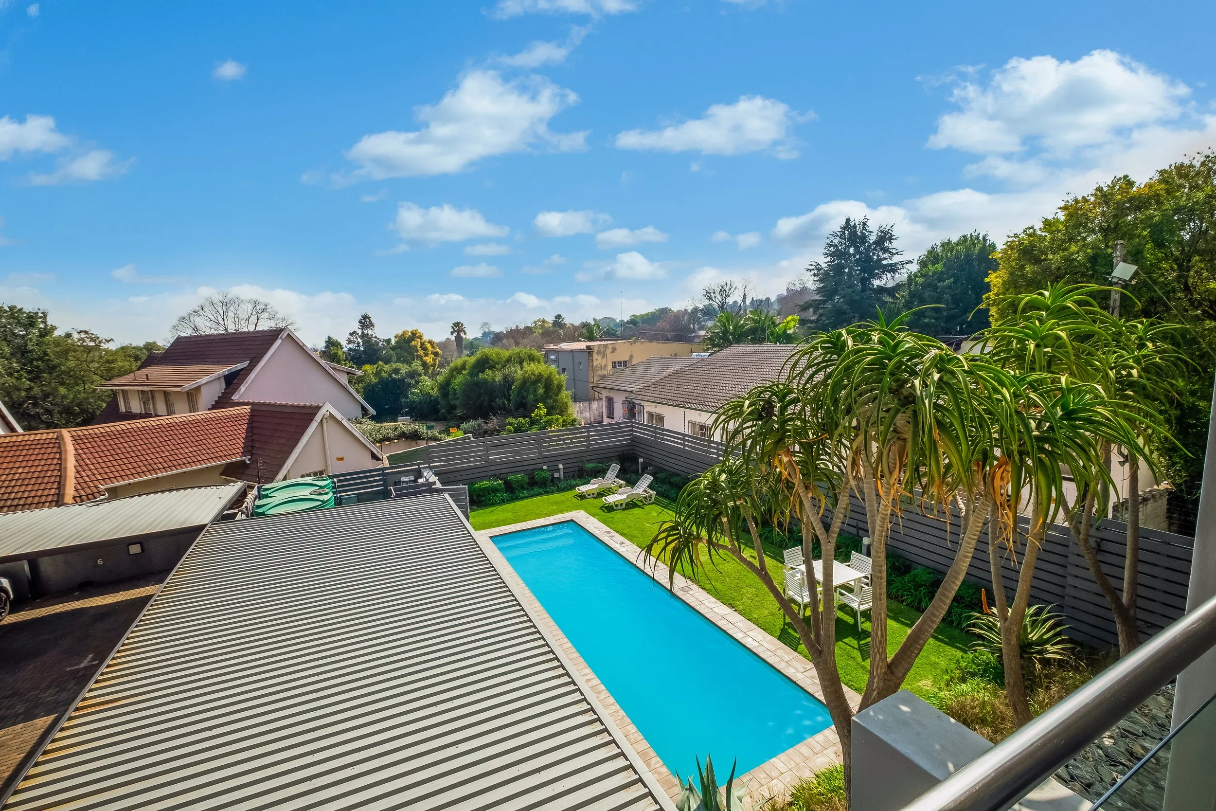 View of a backyard with a swimming pool, lounge chairs, trees, houses, and a bright sky with scattered clouds.