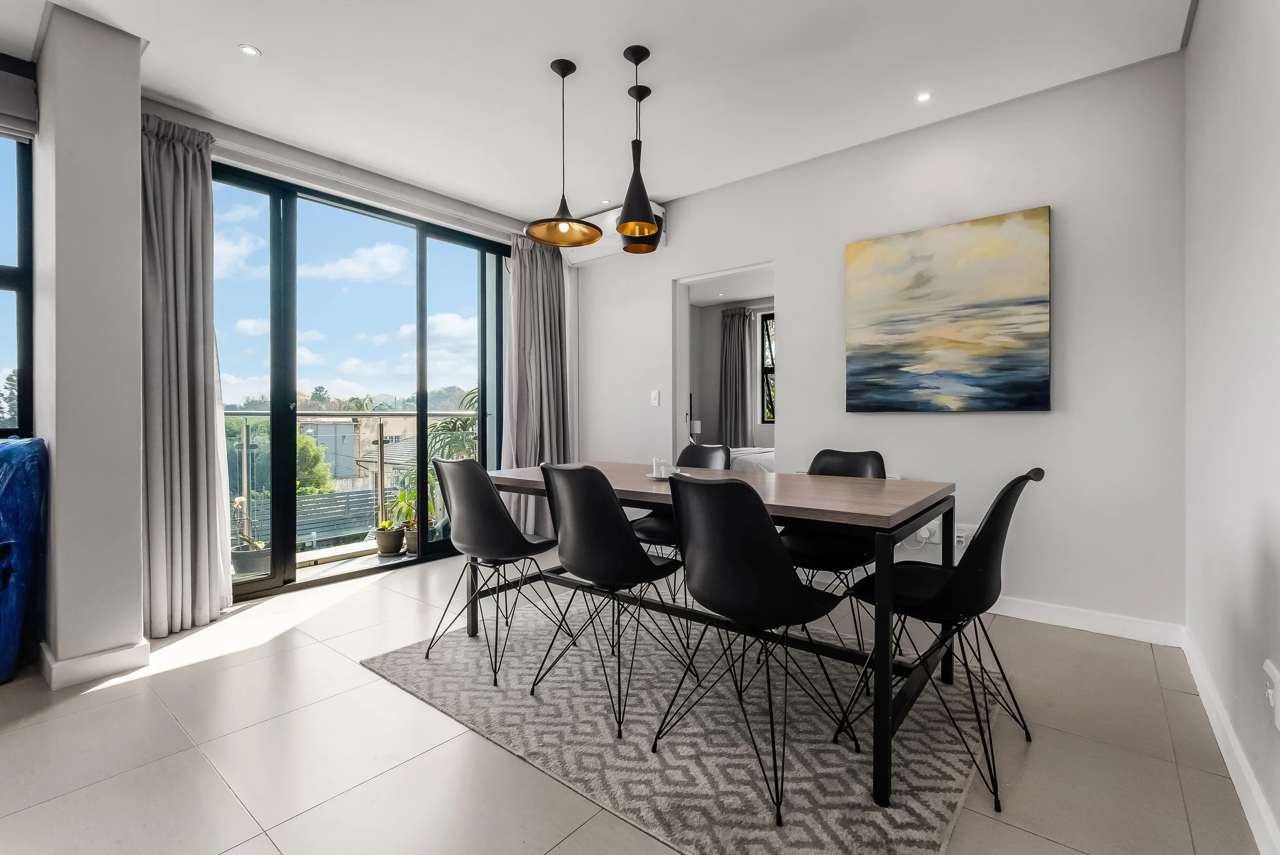 Modern dining room with black chairs, a wooden table, a gray patterned rug, large sliding glass doors with white curtains, a landscape painting on the wall, and a view of trees and houses outside.