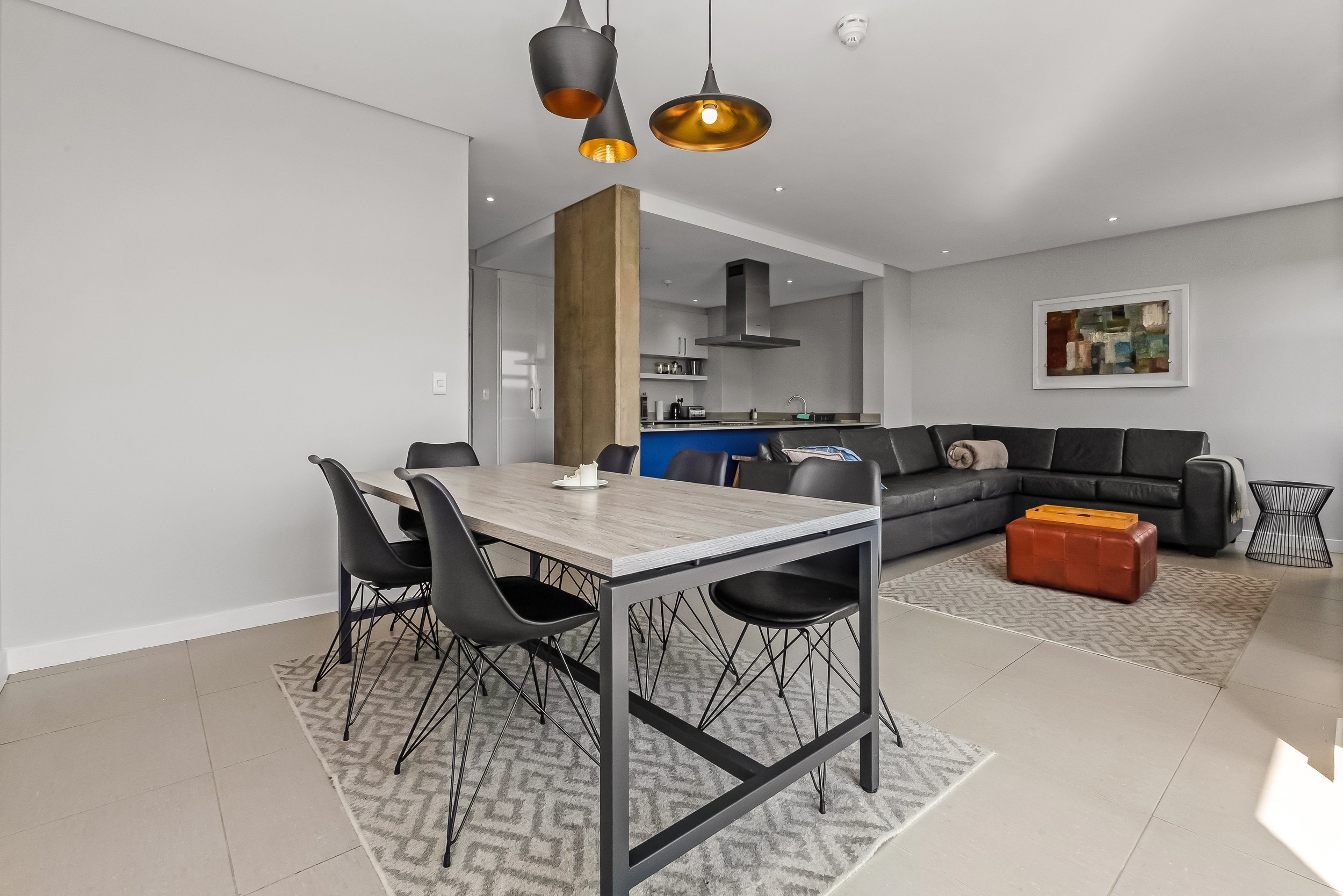 Open-plan living and dining area with a dining table, black chairs, a gray sofa with a blanket, a colorful ottoman, wall art, modern hanging light fixtures, and a kitchen with stainless steel appliances and open shelving.