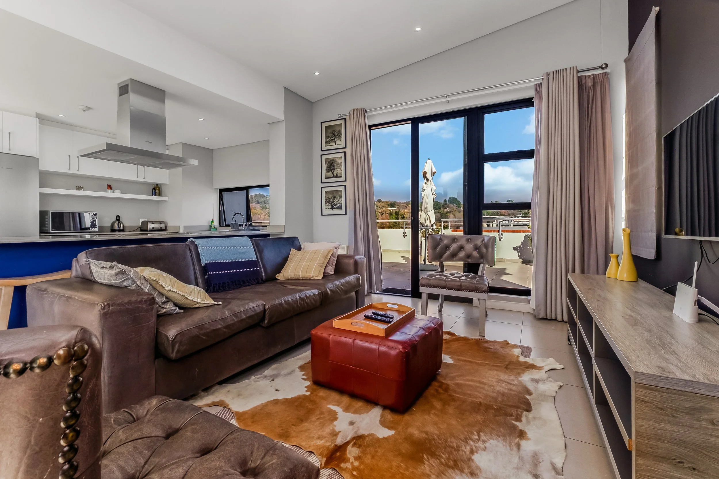 Living room with brown leather sofa, cowhide rug, red leather ottoman, and sliding glass door leading to balcony with outdoor furniture and mountain view.