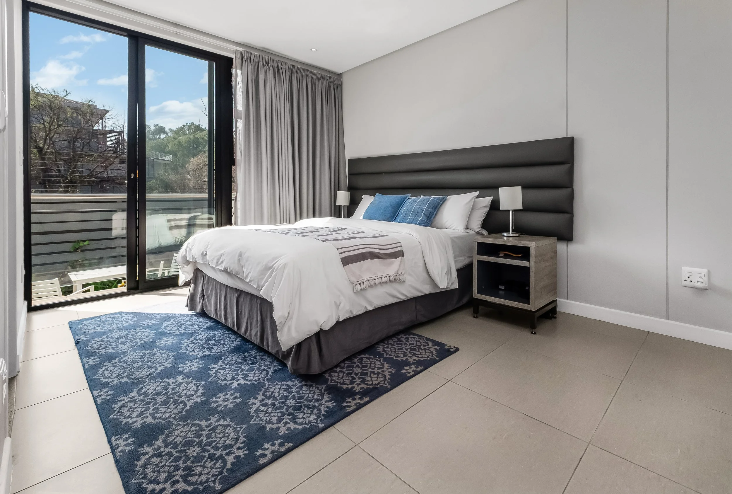 Modern bedroom with large sliding glass door leading to balcony, white walls, grey headboard, bed with white and grey bedding, blue pillows, small bedside table with lamp, blue patterned rug, outside trees and blue sky visible.