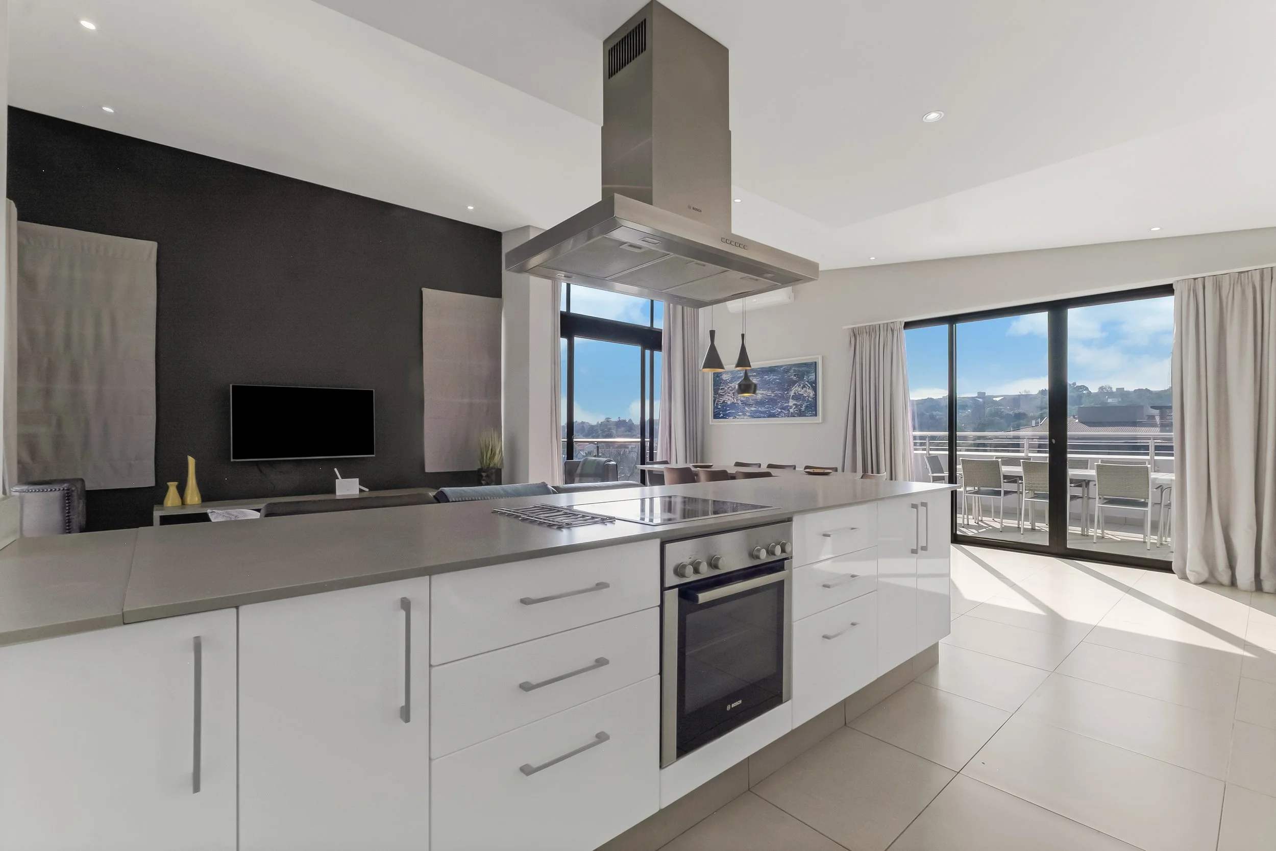Modern kitchen with white island, stainless steel range hood, and large sliding glass doors leading to a balcony with outdoor seating. The room has a black accent wall, a wall-mounted television, and window curtains. There are city and hillside views outside.
