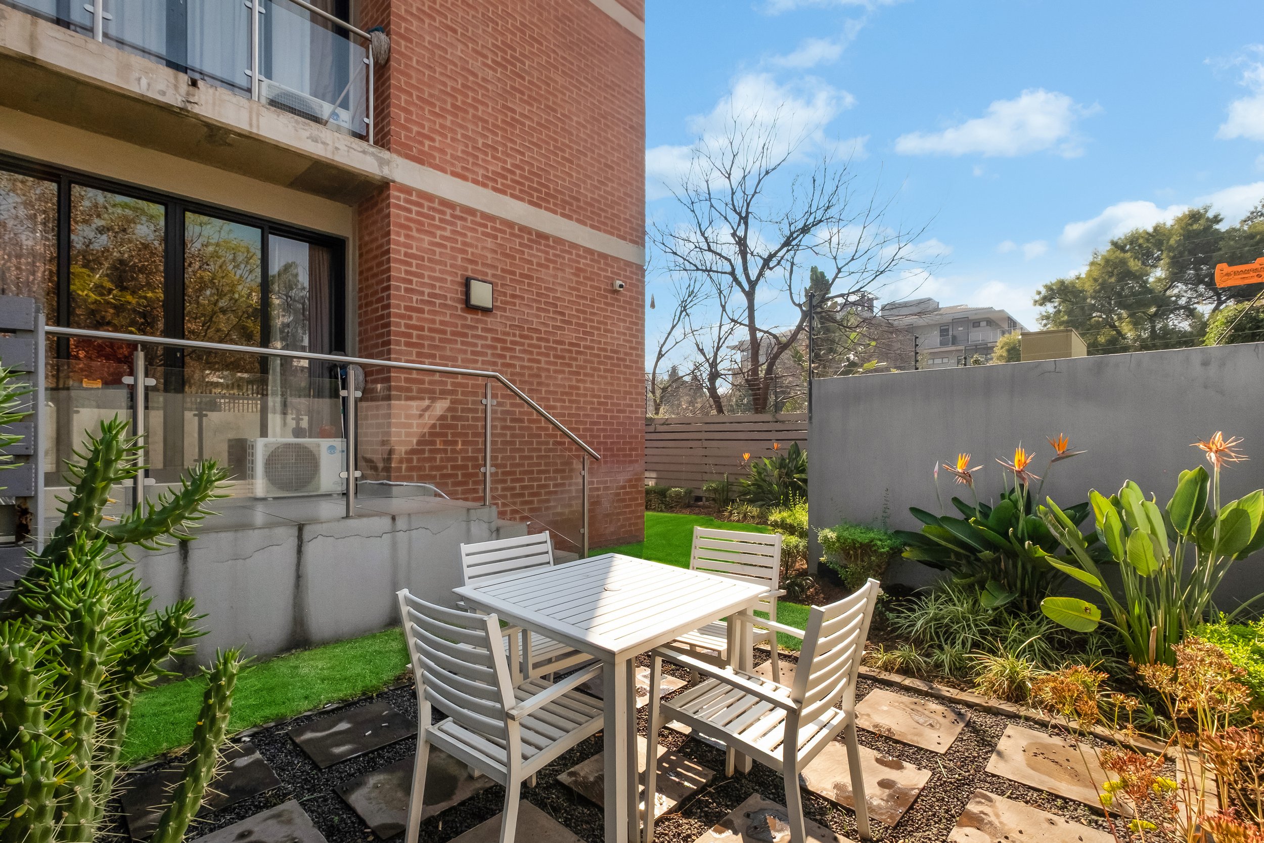 A small outdoor patio area with a white wooden table and matching chairs, surrounded by various green plants including succulents and flowering plants with orange flowers. A gray concrete wall separates the patio from neighboring buildings, with some trees and a blue sky with scattered clouds visible in the background.