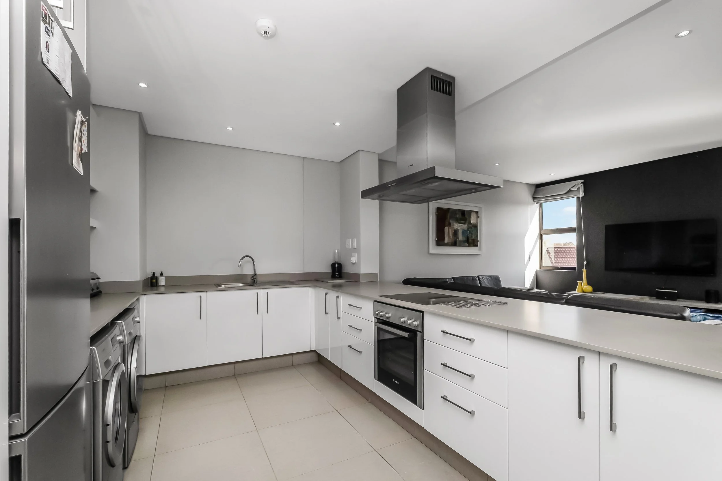 Modern kitchen with white cabinets, stainless steel appliances, gray countertop, black accent wall, and a window with a view of rooftops and blue sky.