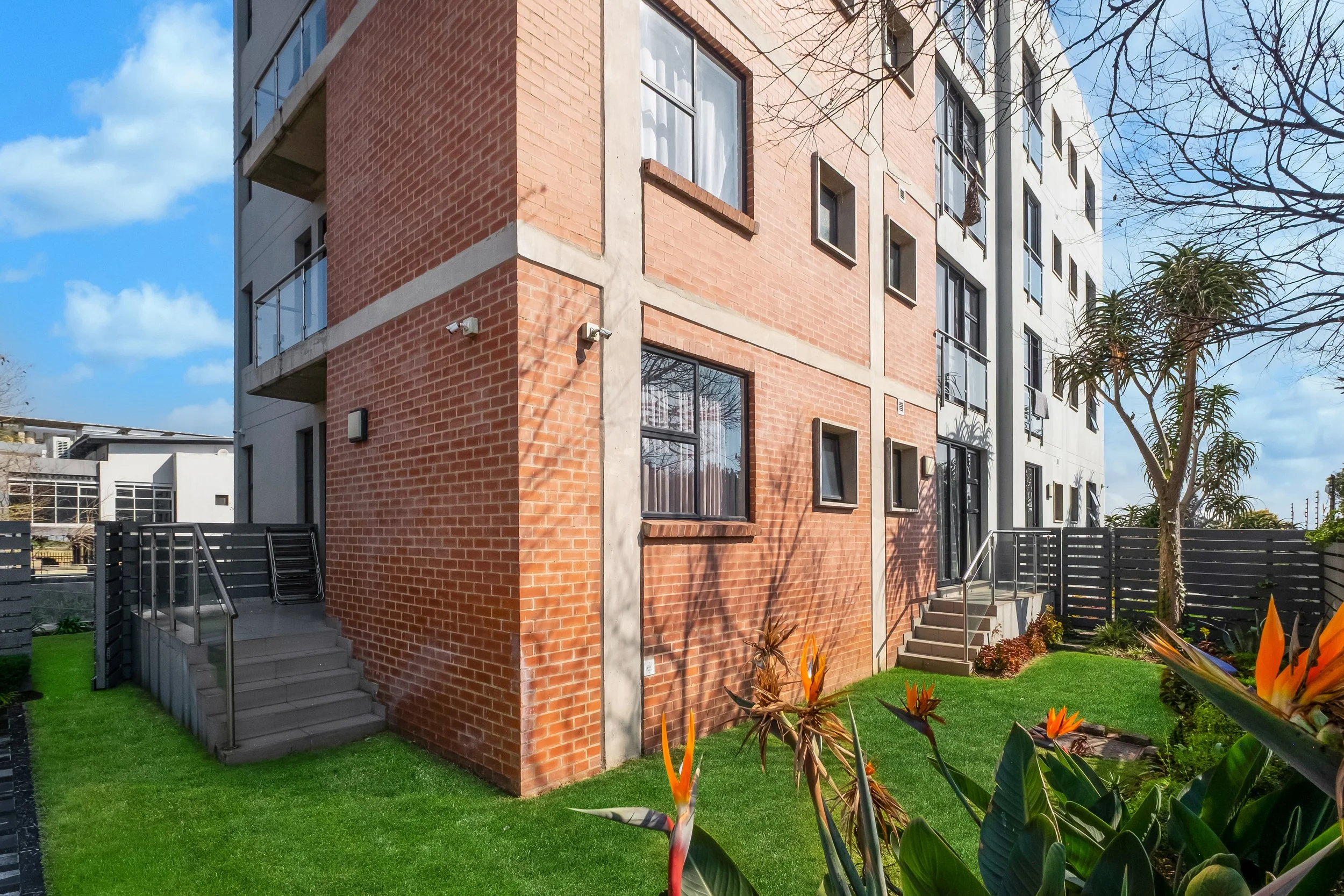 A modern multi-story apartment building with a red brick exterior, large windows, and a concrete and glass balcony. The building is surrounded by a well-maintained green lawn with flowering plants, and a metal staircase leads to a second-floor entrance. The sky is partly cloudy with some trees visible, indicating a suburban setting.