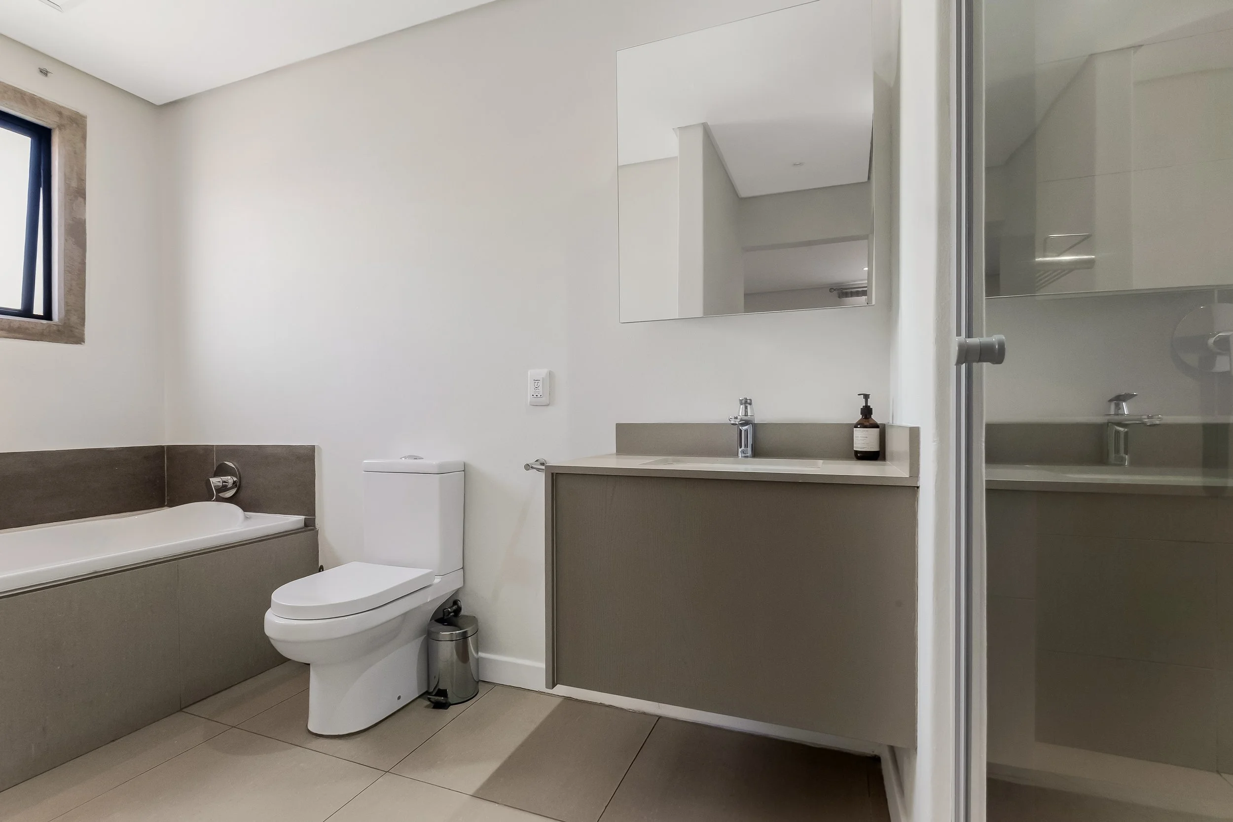 Modern bathroom with a white toilet, bathtub, and a vanity with a sink. A mirror is above the vanity, and there is a small window with a stone frame on the left wall. The bathroom has beige and white tiles.