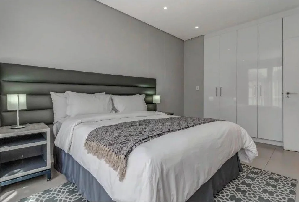 Modern bedroom with a large bed, white bedding, gray headboard, side tables with small lamps, white closet doors, and a gray patterned rug.