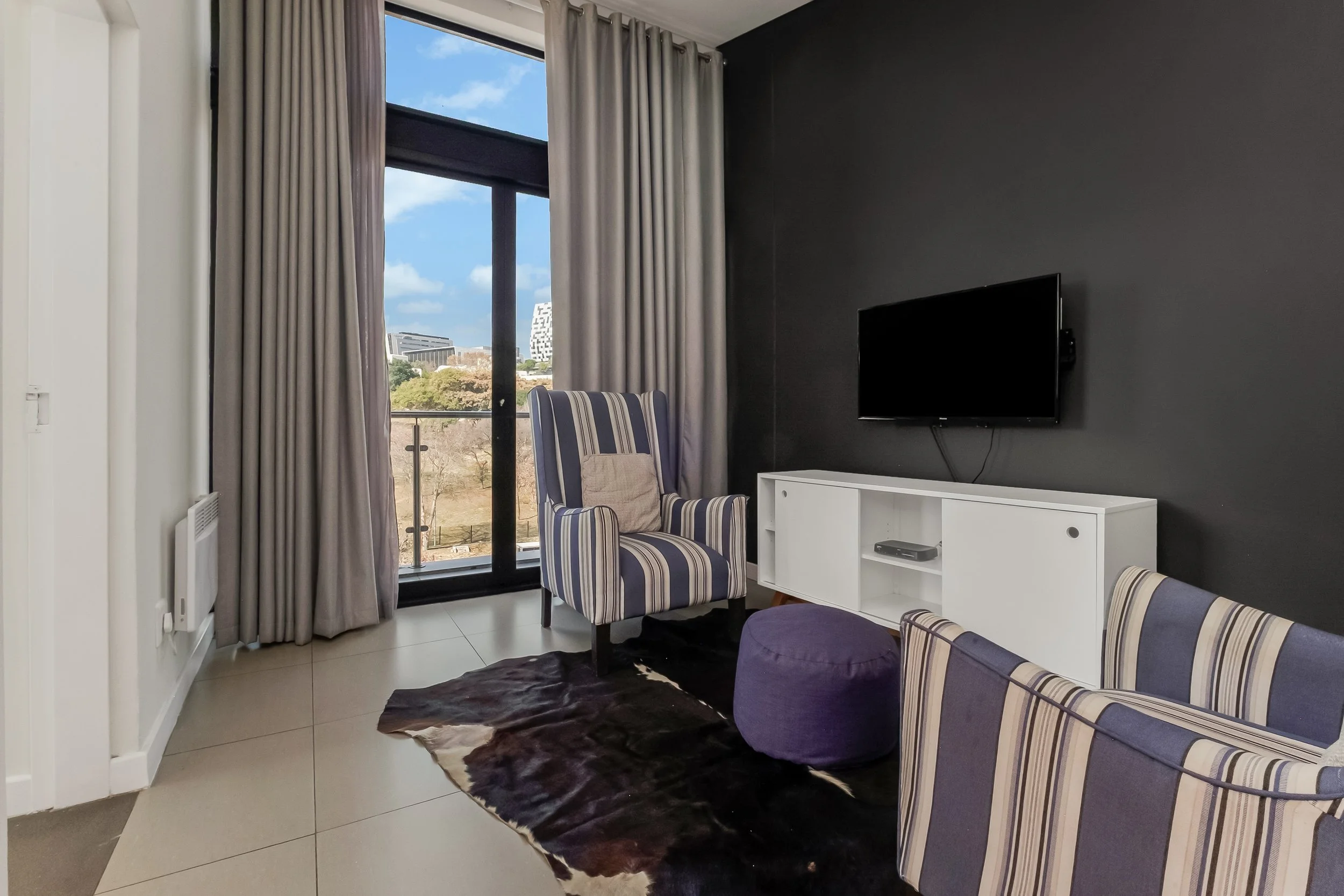 Living room with large window showing blue sky and buildings outside, gray curtains, striped armchair, TV mounted on dark wall, white cabinet with a black device, and cowhide rug on tile floor.