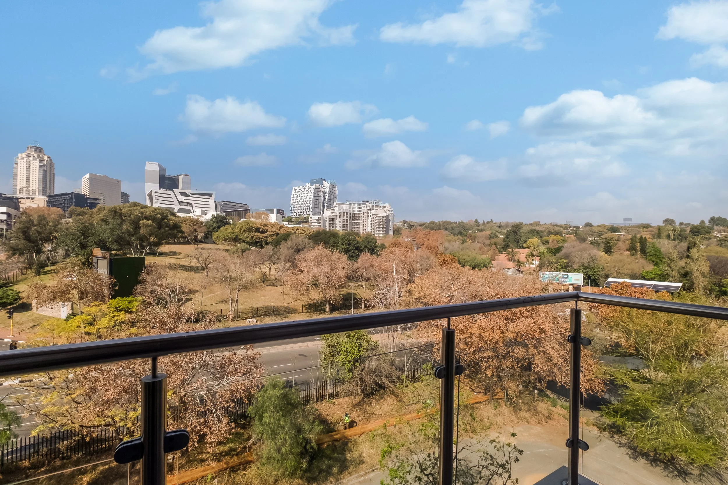 View from a balcony overlooking a city with modern buildings and trees with autumn foliage on a partly cloudy day.