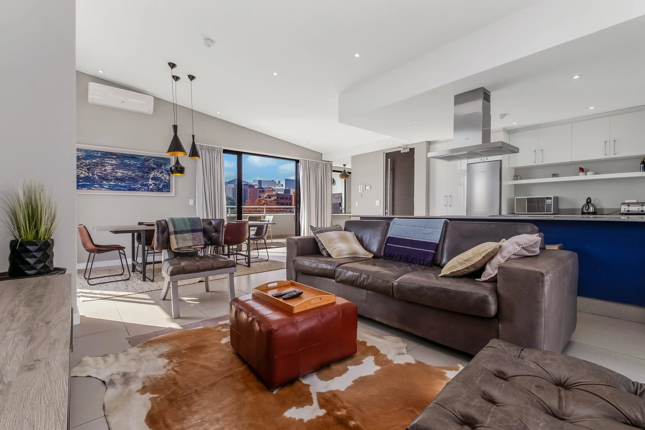 Modern living room with brown leather sofas, a cowhide rug, a small red leather ottoman, a dining area with chairs, a blue and white painting, large windows showing city buildings, and a kitchen with white cabinets and a stainless steel range hood.