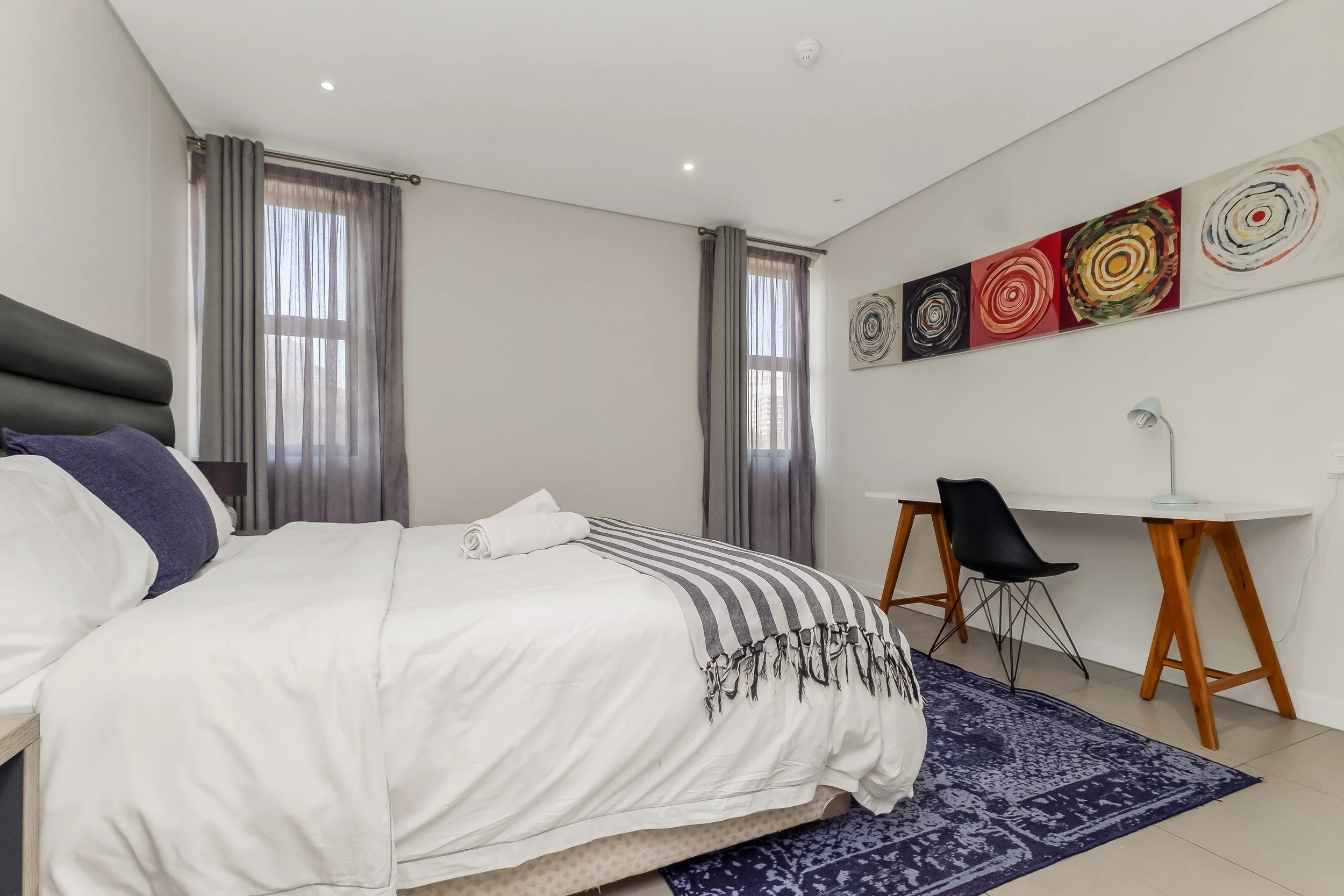 A bedroom with a bed, purple pillows, a striped blanket, two windows with sheer curtains, a modern desk with a black chair, a white desk lamp, a colorful abstract circular art piece on the wall, and a blue patterned rug.