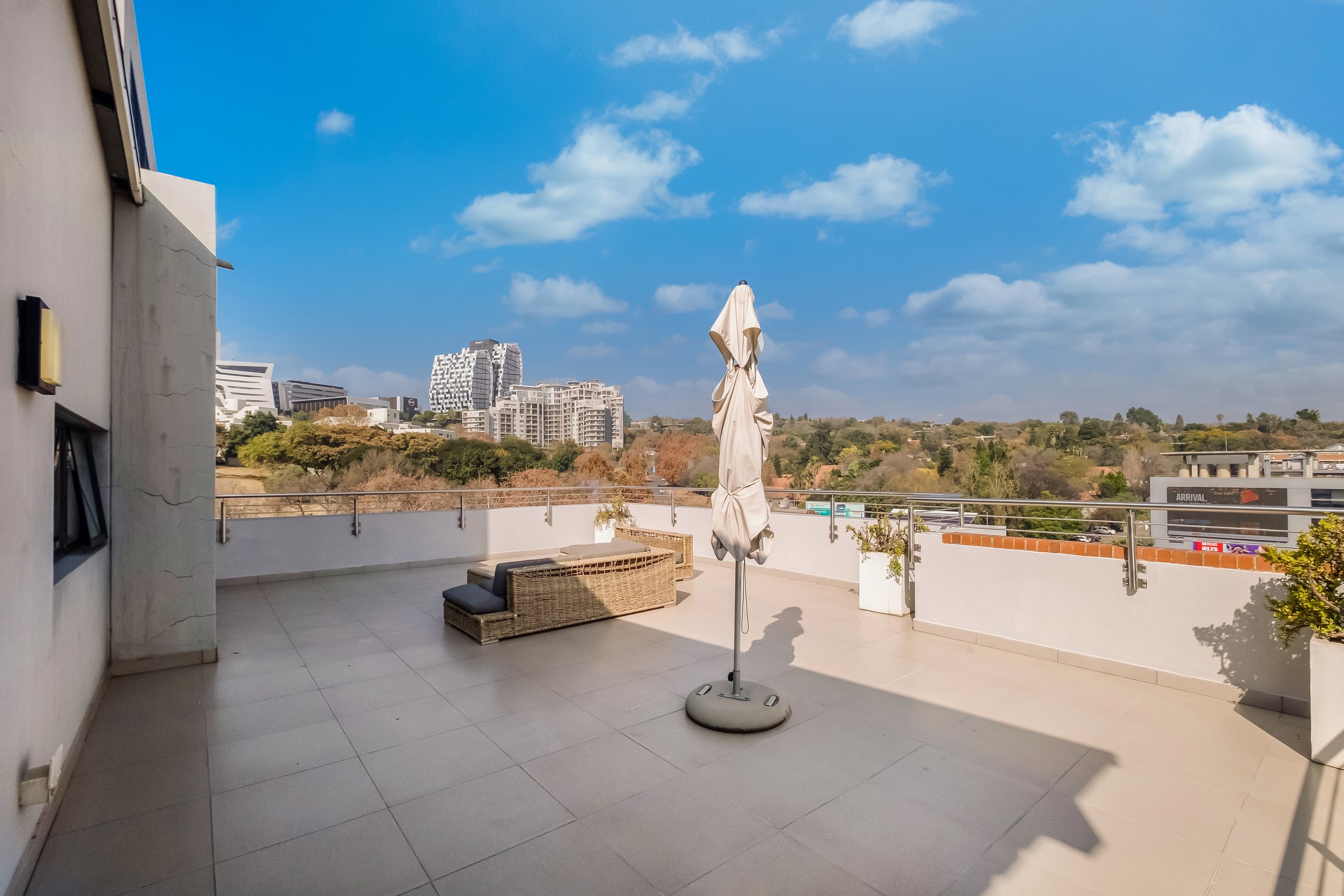 A spacious balcony with beige tiled flooring, a wicker sofa with cushions, a closed beige patio umbrella, and a white railing. The background features a cityscape with modern buildings, greenery, and a bright blue sky with scattered clouds.