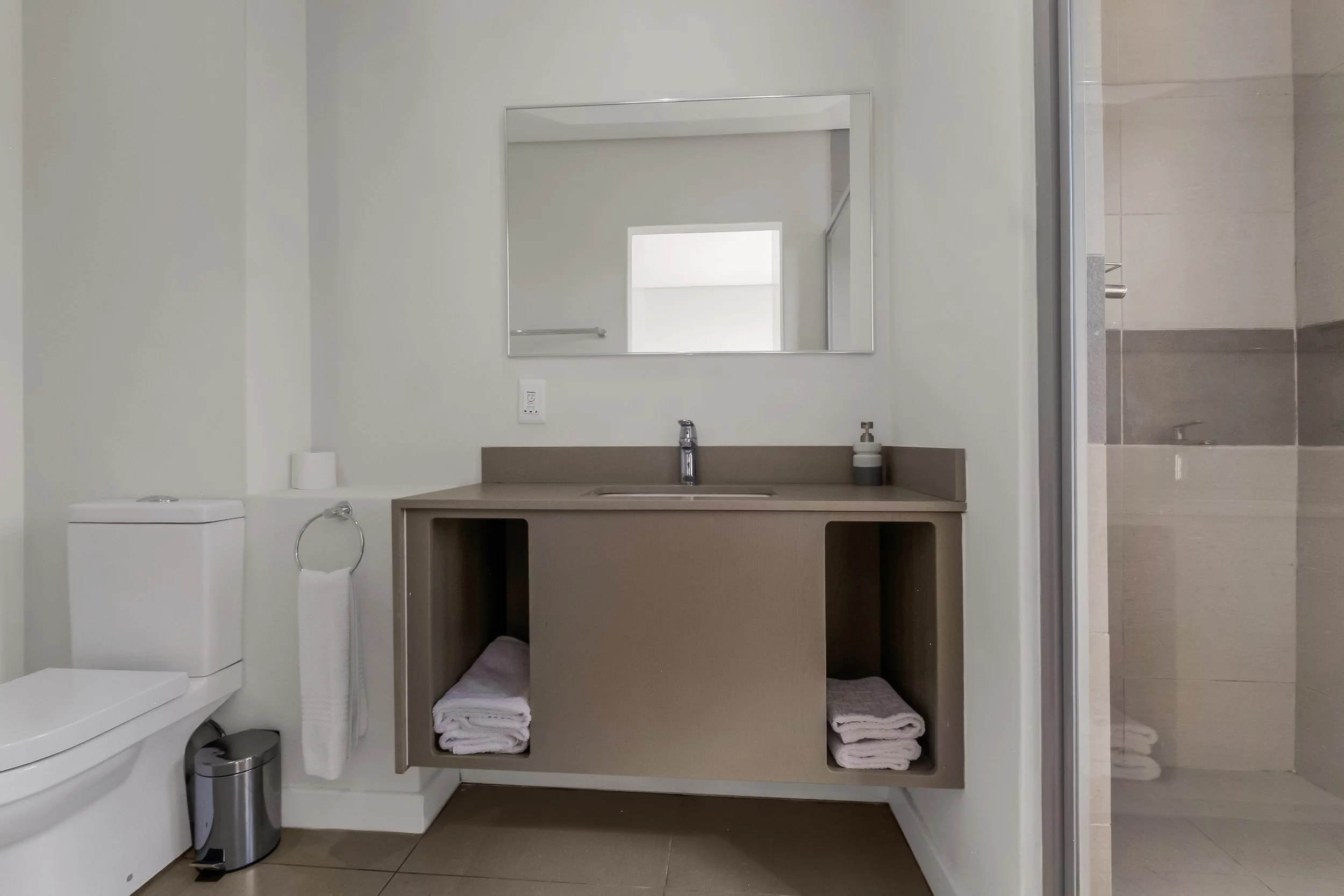 A bathroom with a mirror above the sink, a towel rack, a countertop with a soap dispenser, a toilet, and a shower with a glass door, and neatly folded towels underneath the sink.