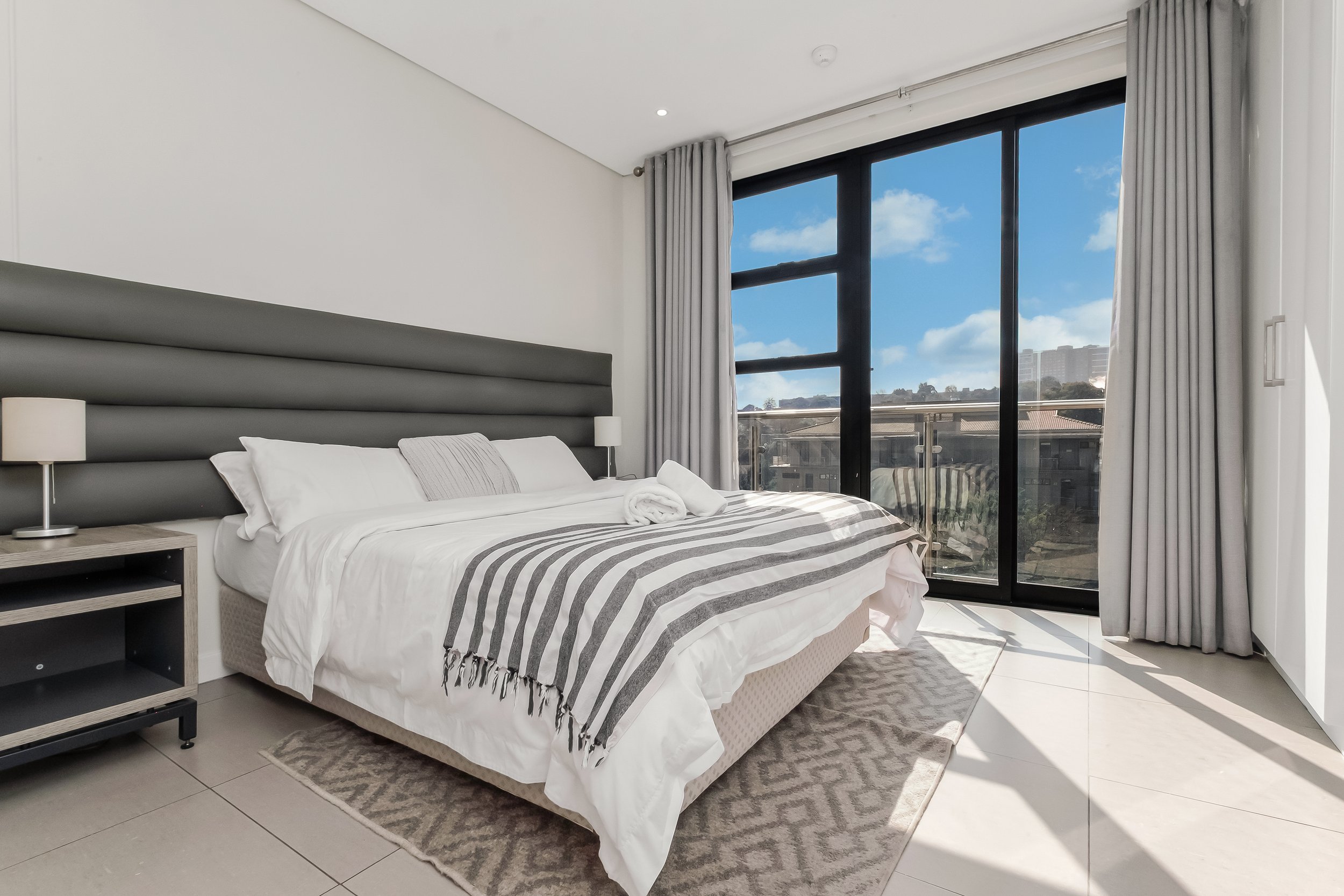 A modern bedroom with a bed featuring a striped gray and white bedspread and white pillows. A nightstand with a lamp is beside the bed. Large sliding glass doors with white curtains reveal a city view and blue sky outside.