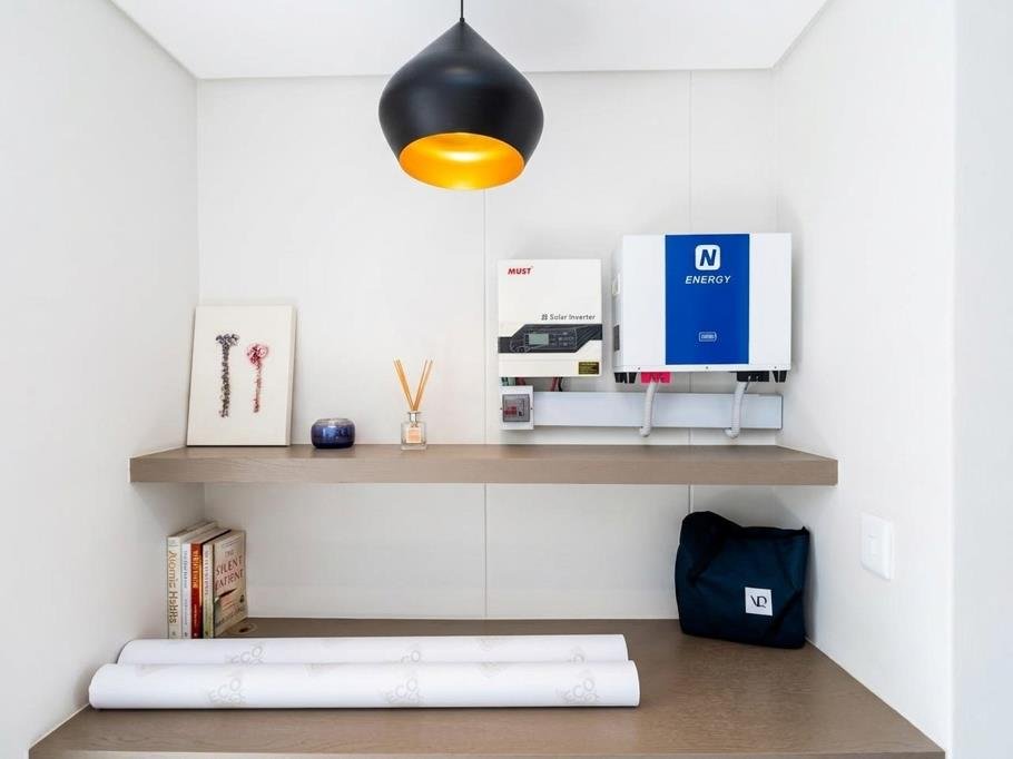 Corner of a room with a floating shelf holding decorative items, books, and an energy system. There is a black pendant light with an orange interior hanging from the ceiling and two rolled-up documents on the table below.