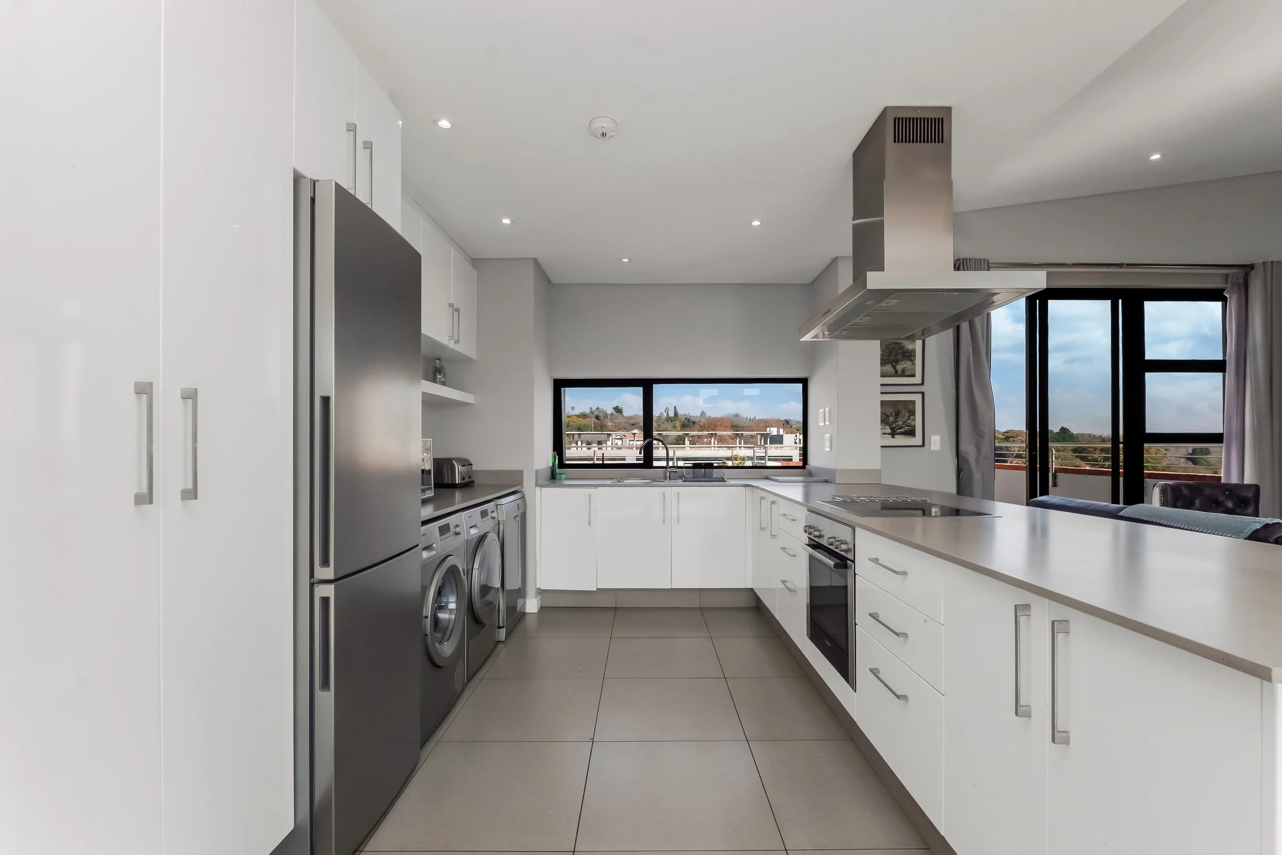 Modern kitchen with white cabinets, stainless steel appliances, and a large window with a view of trees and buildings outside.