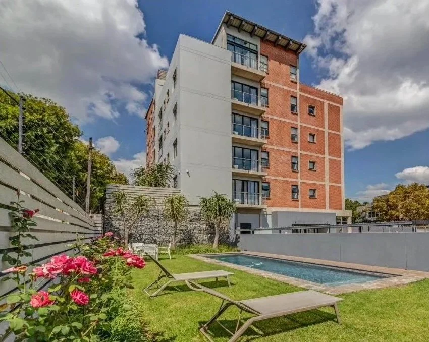 Outdoor view of a modern apartment building with balconies, a backyard with a swimming pool, lounge chairs, pink flowers, and trees under a partly cloudy sky.