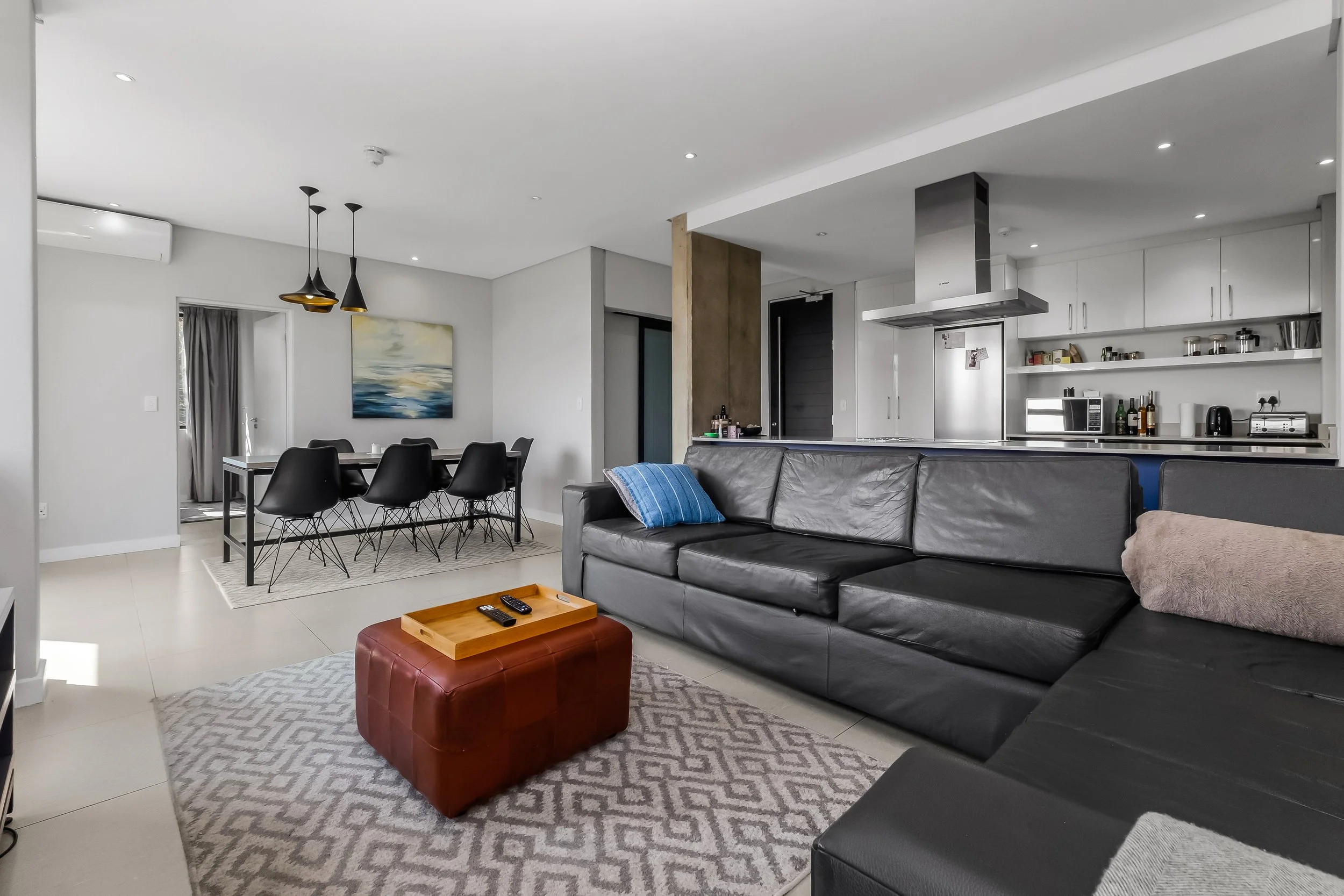 Living room with black sofa, small red ottoman, and wooden tray with remote controls, dining area with black chairs, artwork on white wall, and open kitchen with stainless steel appliances.