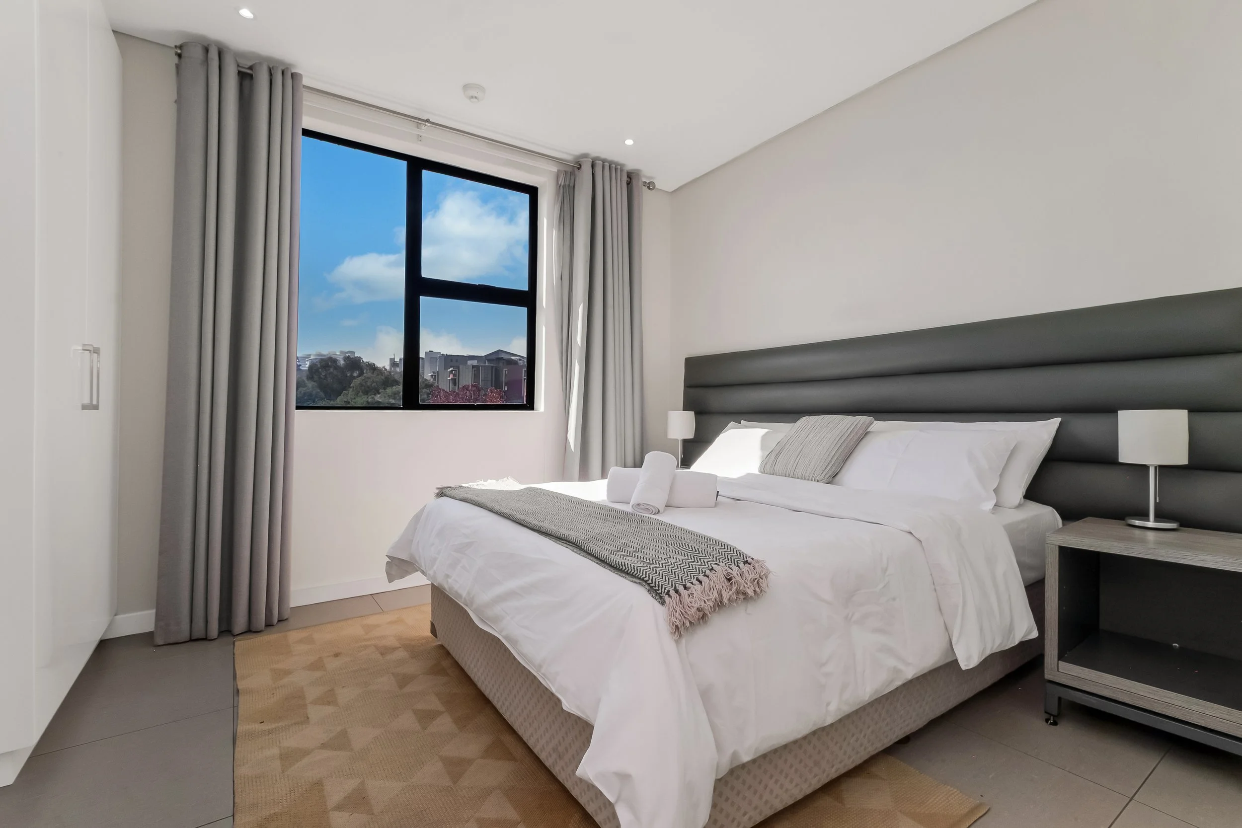 Modern bedroom with a large window showing a blue sky and buildings outside, featuring a bed with white linens and rolled towels, a gray headboard, bedside lamps, and light wood flooring.
