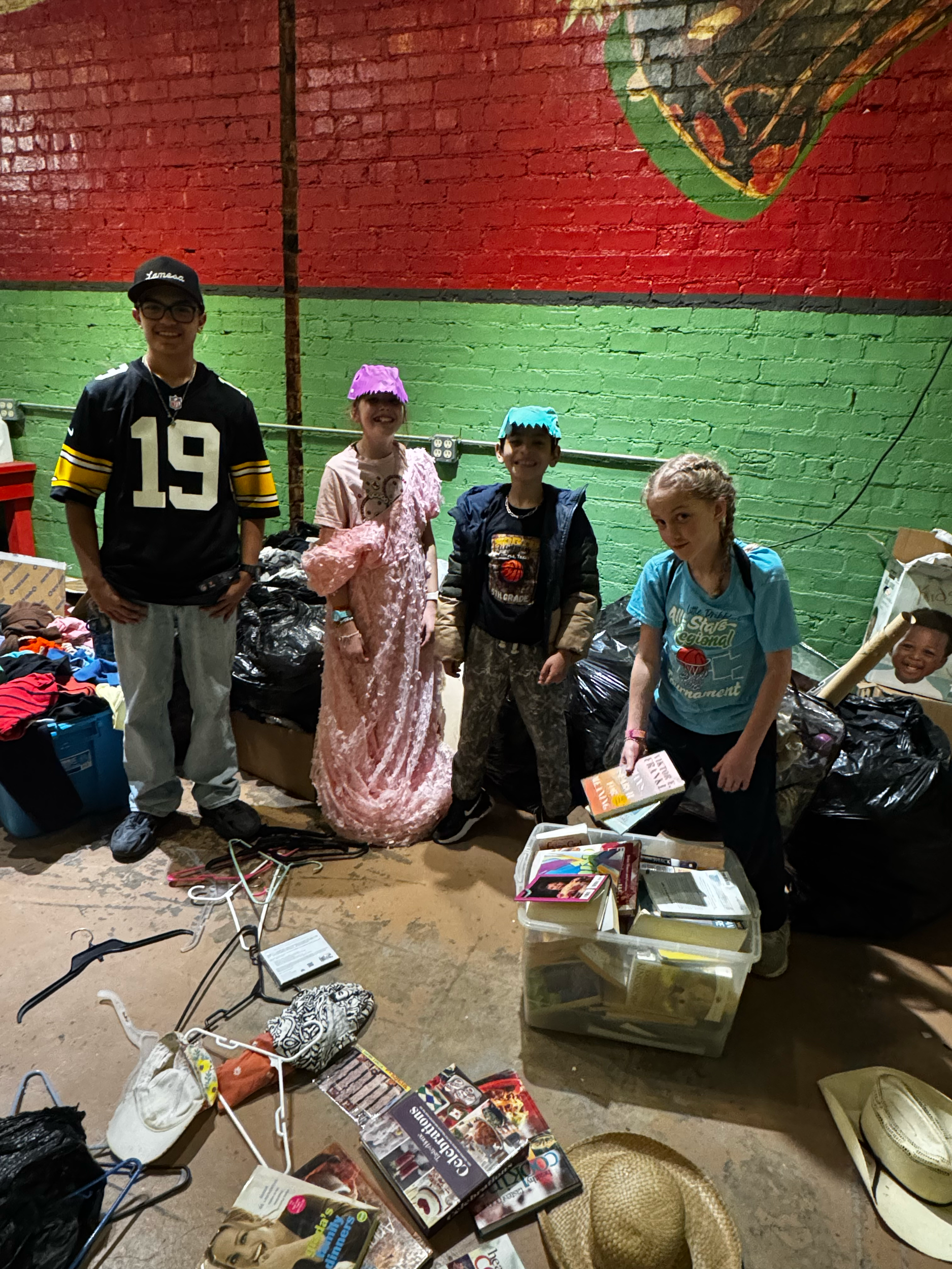 Four children standing together in a basement or garage with a red and green painted brick wall and various piles of clothes, magazines, and items on the floor around them.