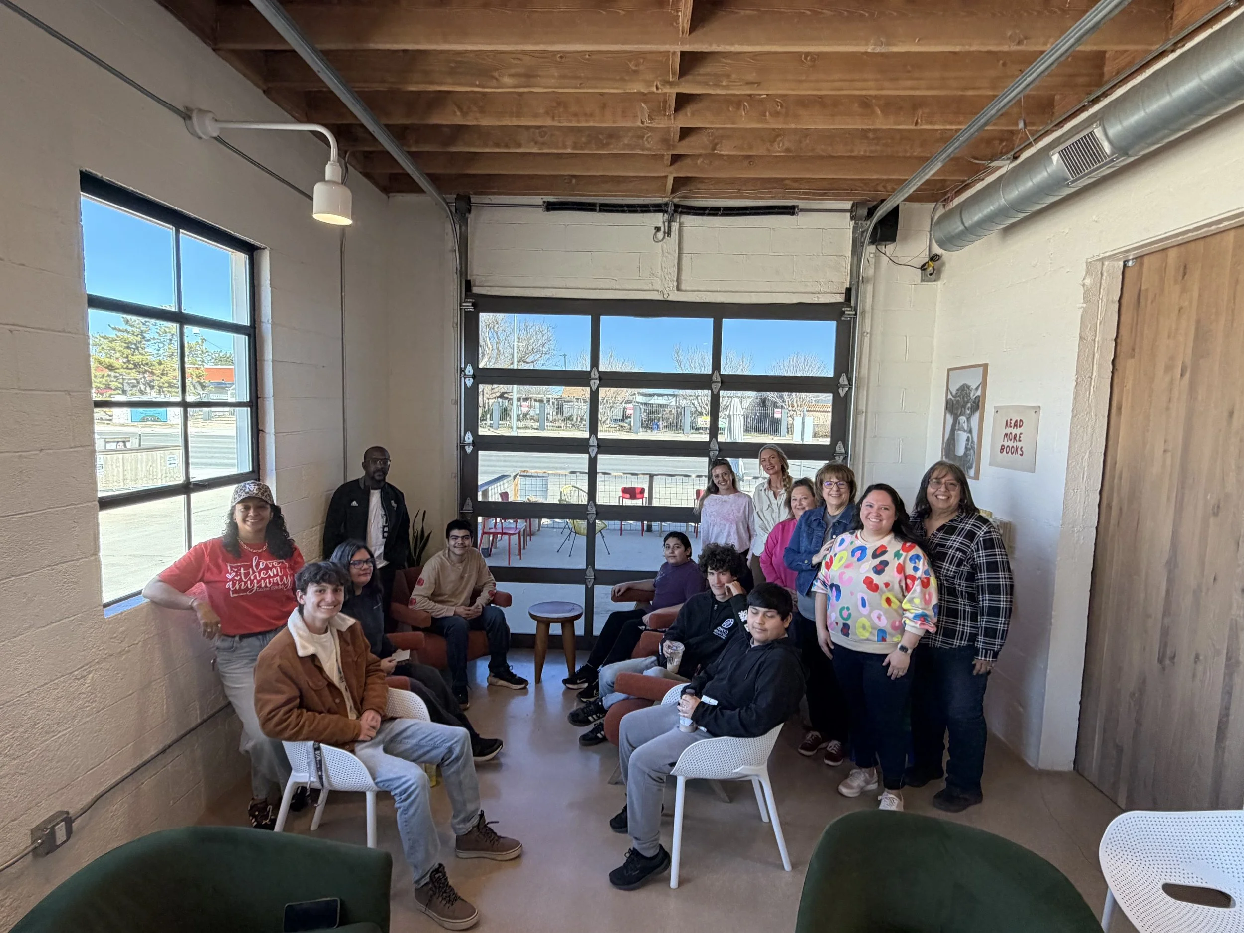 A group of diverse people gathered indoors, some sitting and some standing, smiling for the camera in a well-lit room with large windows and an industrial-style ceiling.
