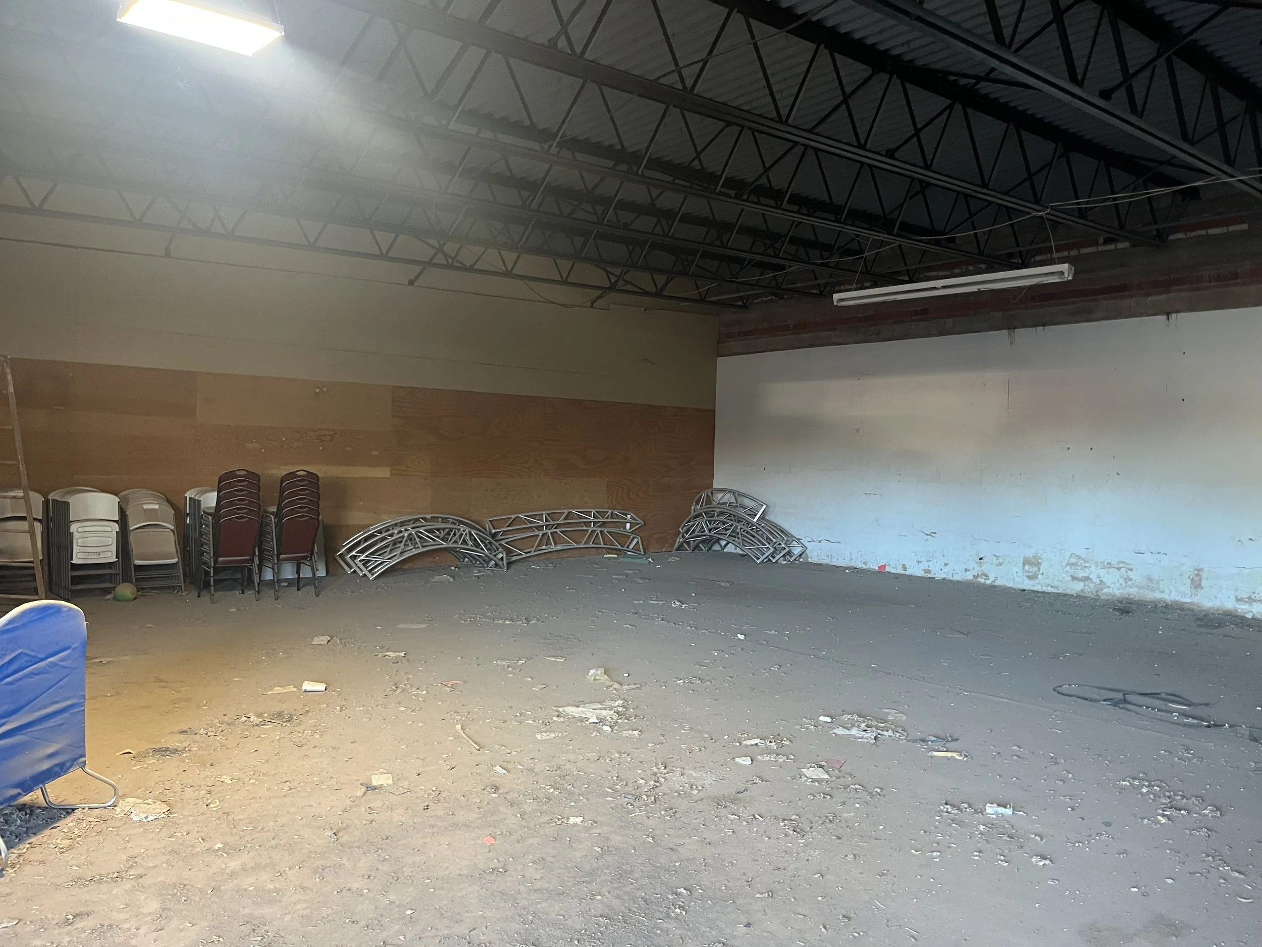 An indoor space under construction or renovation with stacked chairs on the left and metal trusses and a light fixture on the ceiling.
