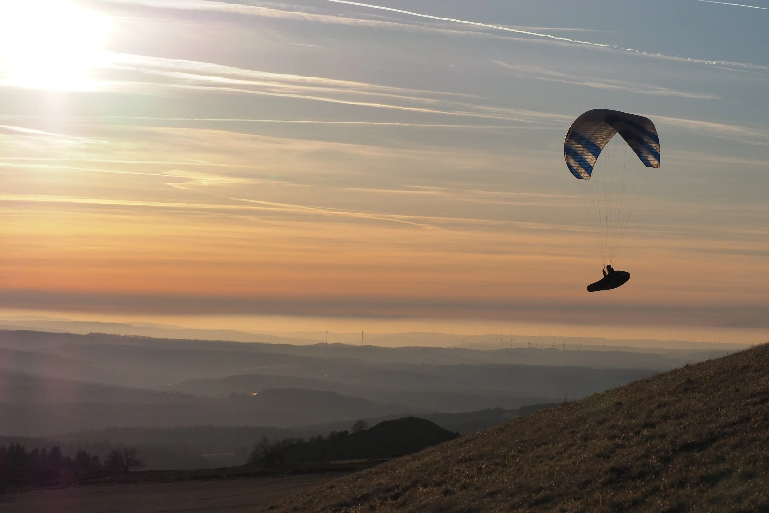 Ein Paragleiter fliegt bei Sonnenuntergang über eine ländliche Landschaft mit Hügeln und Wäldern.
