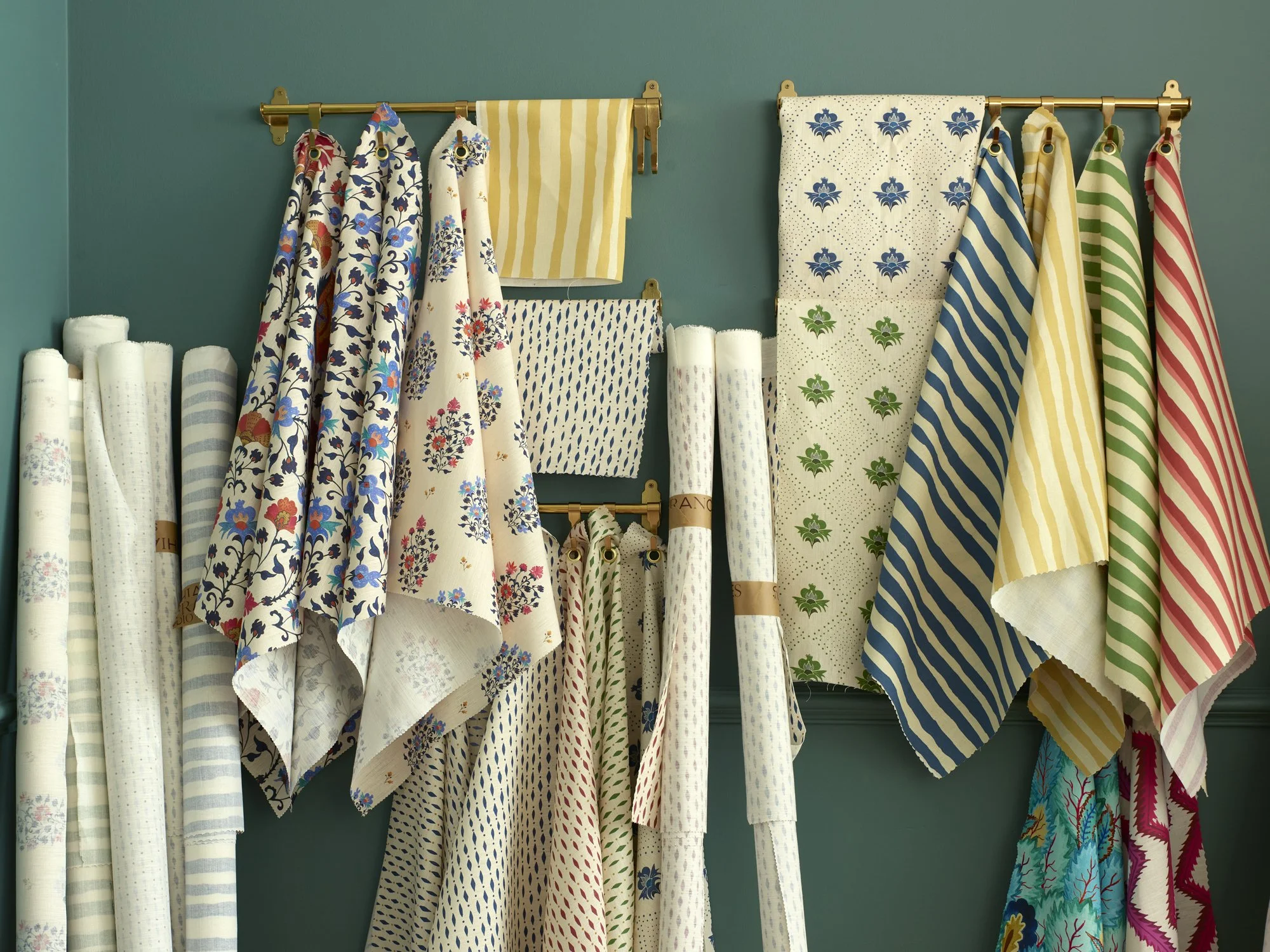 Display of various patterned fabrics hanging on rods against a teal wall, with some rolls of fabric on the floor.