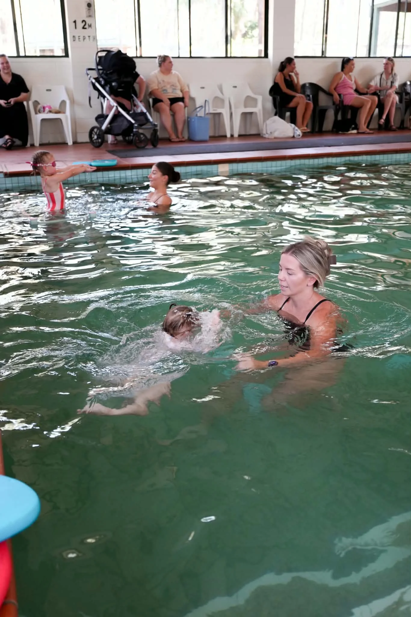 A swimming pool scene with children and adults. A girl in a red and white swimsuit is being held by a woman in the pool. Several people are sitting on chairs along the poolside, some talking, some watching the activity in the pool. There is a stroller and bags beside the seating area. Large windows in the background let in natural light.