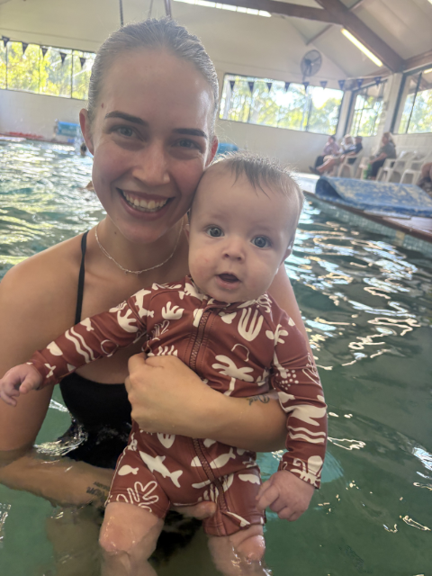 A woman and a young girl in a swimming pool, smiling and enjoying their time together. The woman is supporting the girl, who is wearing pink goggles and a striped swimsuit. The background shows poolside equipment and colored towels.