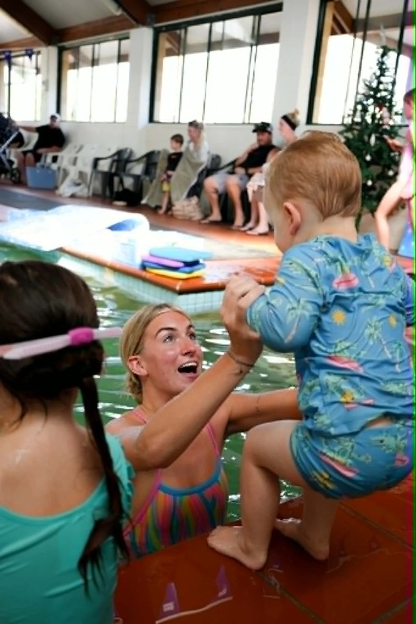 A woman is in a swimming pool holding a young boy dressed in a blue floral swimsuit. Two other girls with long hair and sunglasses are also in the pool, one with glasses and a striped swimsuit, and the other with a turquoise swimsuit and a pink headband. They appear to be enjoying a sunny day indoors, with a Christmas tree visible in the background.
