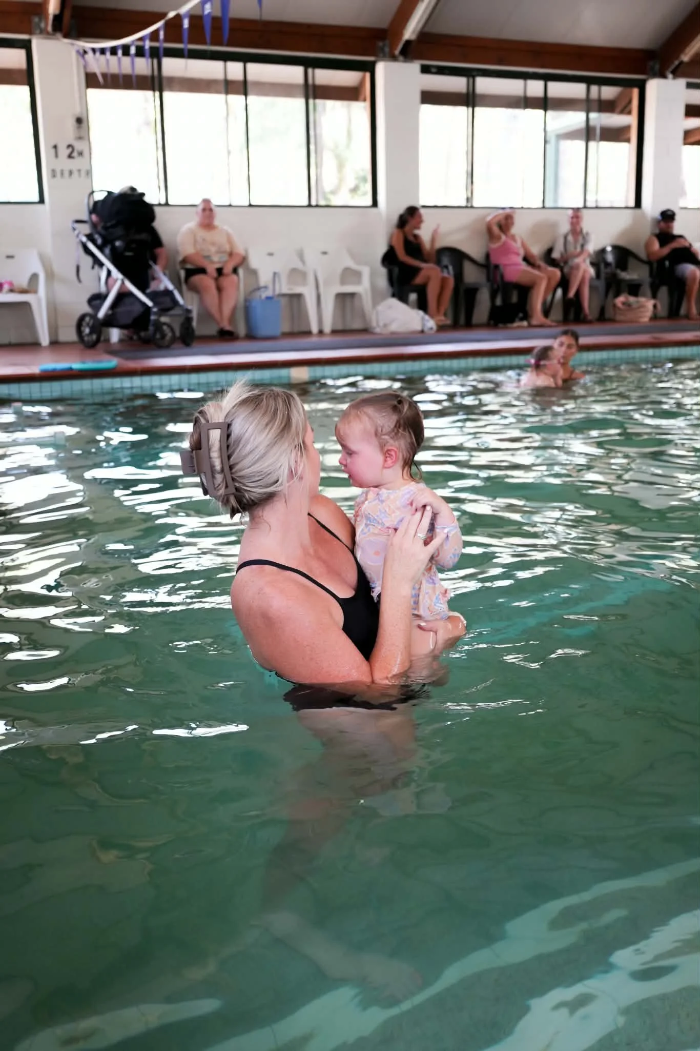 A woman holding a young child in a swimming pool with others sitting on chairs along the back wall of the pool area.