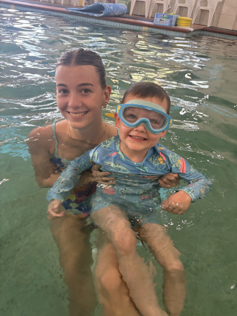 A young girl wearing pink swimming goggles and a blue swimsuit with floral pattern, playing in a swimming pool, splashing water with her mouth open, with water droplets around her and a blurred background of greenery and pool surroundings.