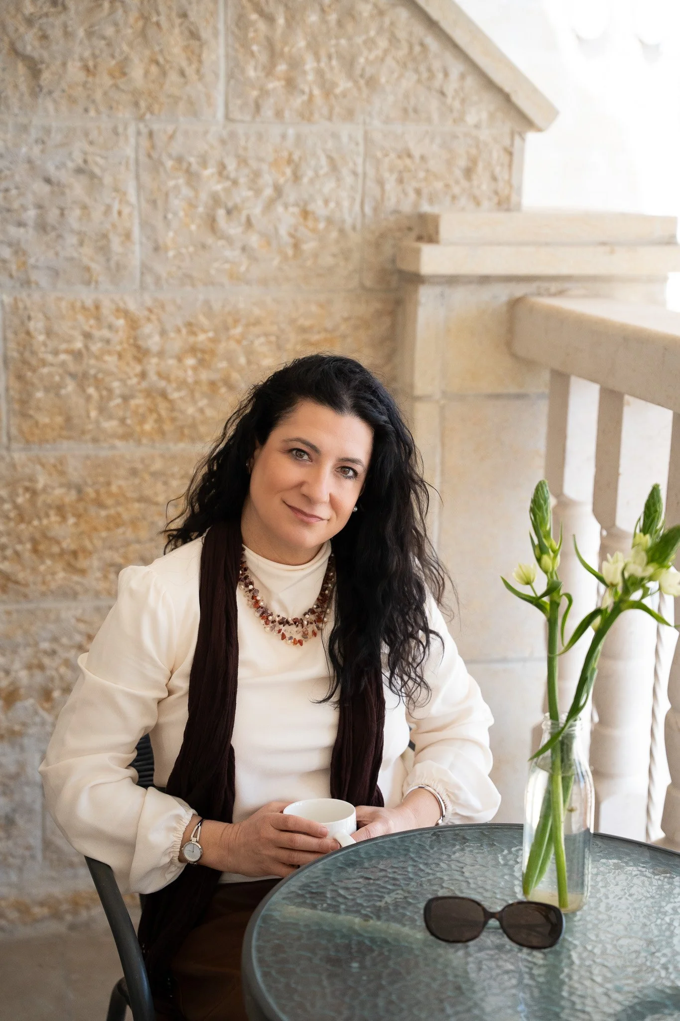 A woman with long dark hair, wearing a cream-colored blouse and brown vest, sitting at a glass table with a vase of white flowers and sunglasses, holding a white mug, in front of a beige stone wall and stair railing.