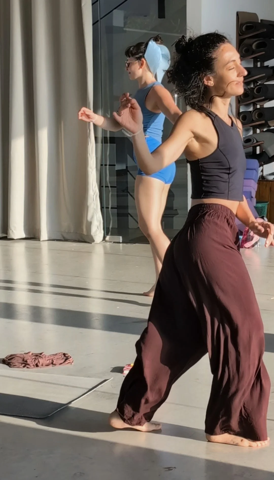 Two women practicing yoga indoors, one in the foreground with dark curly hair wearing a black sports top and loose brown pants, and the other in the background with dark hair tied back wearing a blue tank top and matching shorts, standing near a glass wall with yoga mats and equipment nearby.