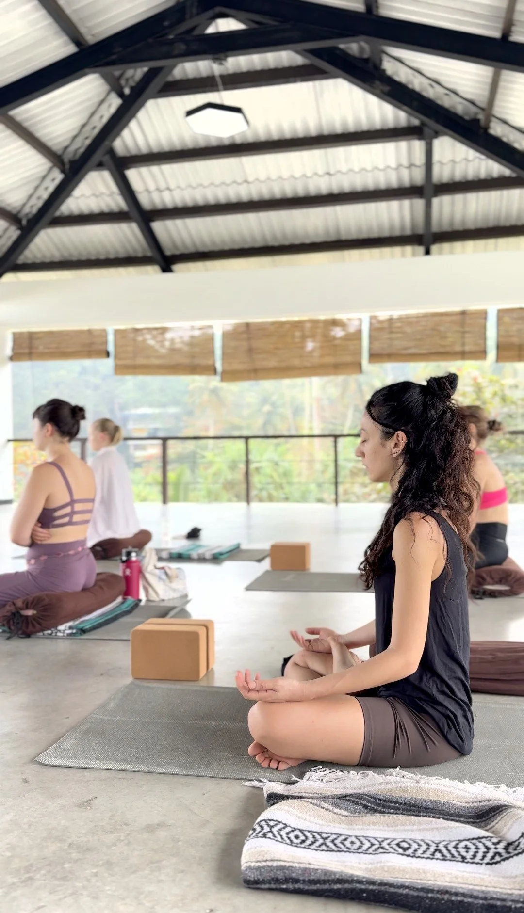 People practicing yoga or meditation in a spacious, well-lit room with large windows, mats, and yoga props.