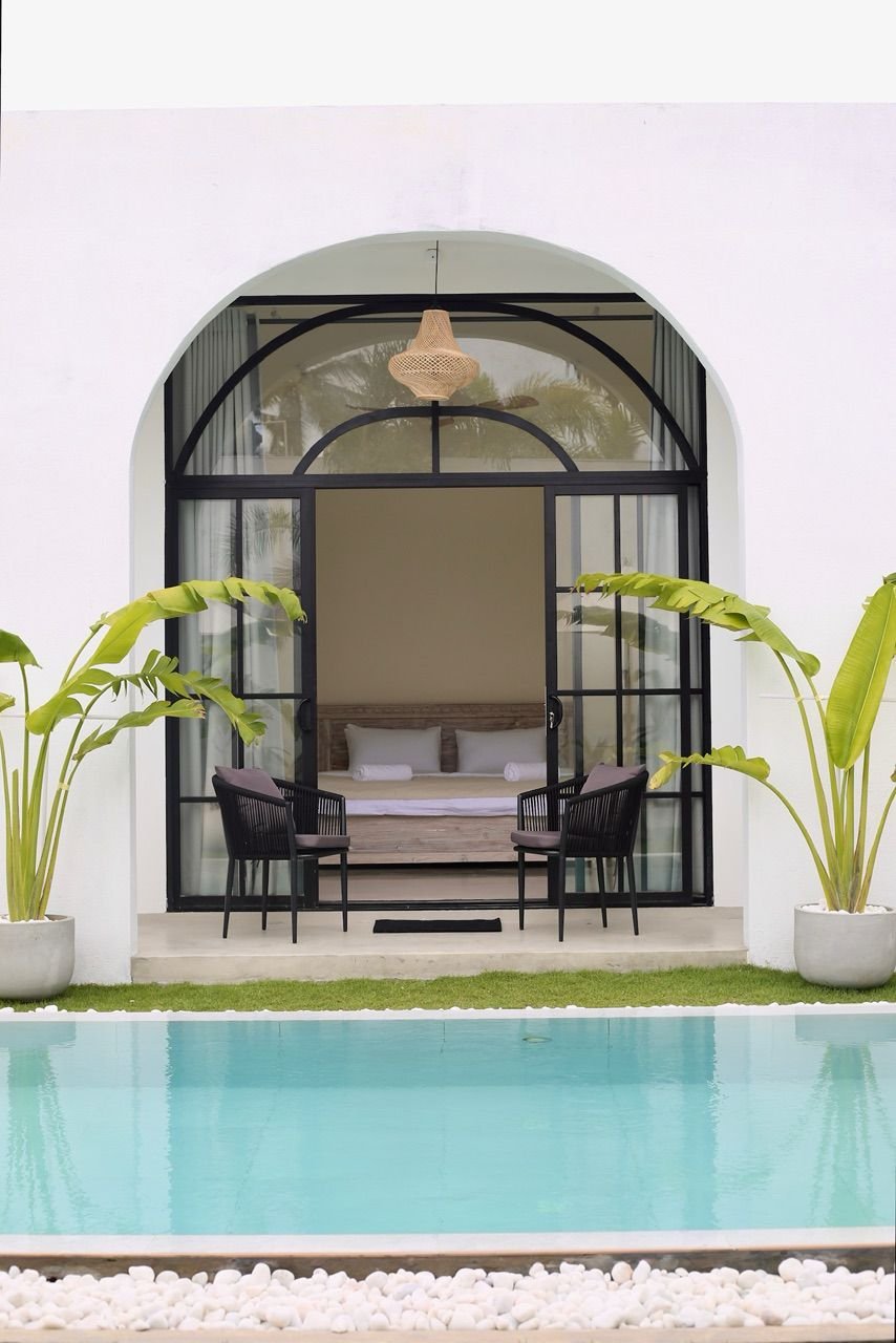 A modern outdoor patio with a view of a swimming pool, large potted banana plants, and a lounge area with a bed inside a glass enclosure.