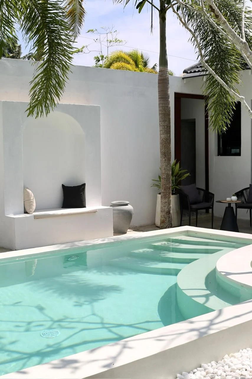 Outdoor pool area with white walls, black and white cushions on a built-in bench, palm trees, and outdoor seating with a small table.