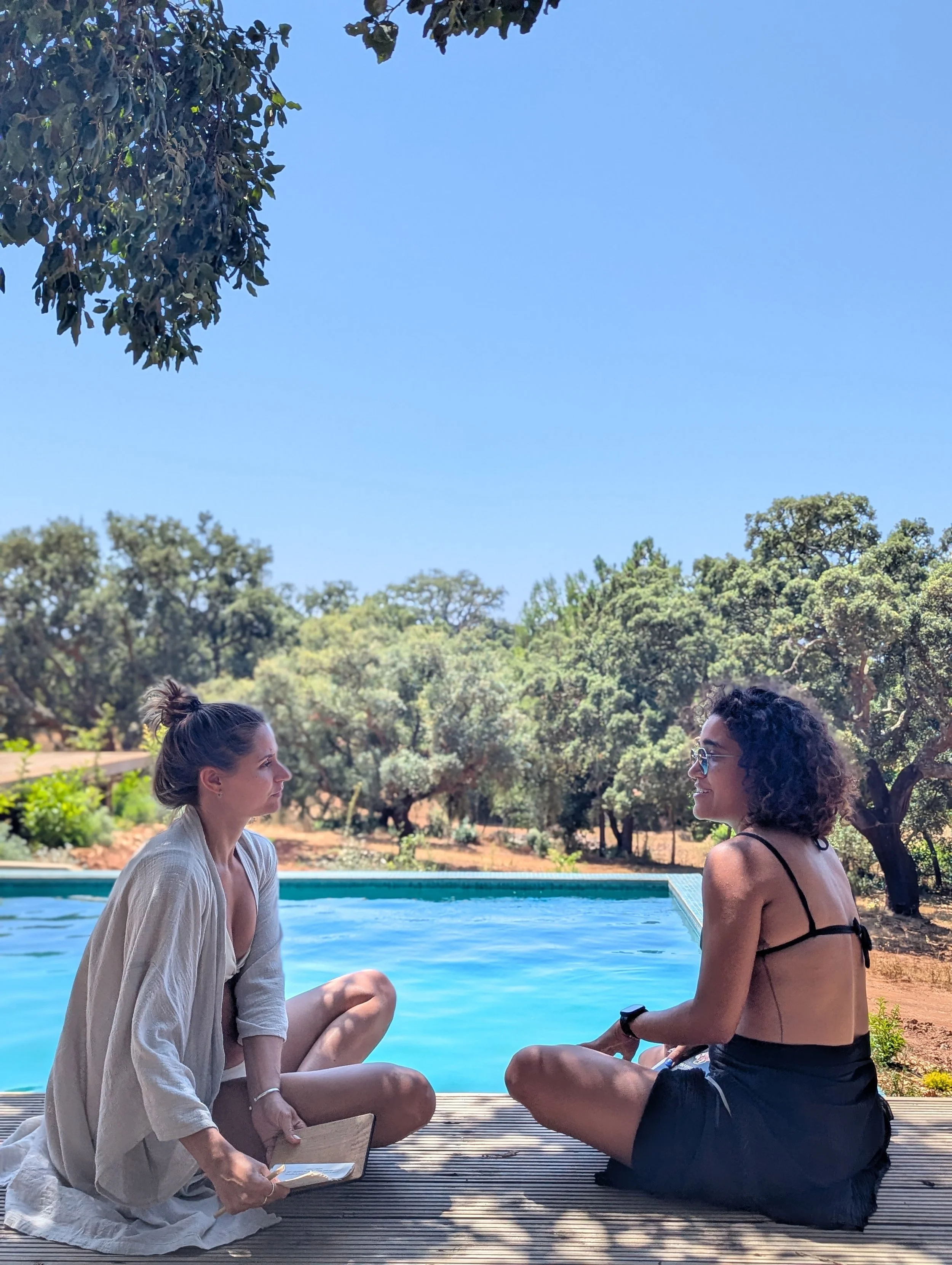 Two women with dark and light hair, sitting cross-legged on a wooden deck near a swimming pool, facing each other and holding hands, with a backdrop of trees and a clear blue sky.