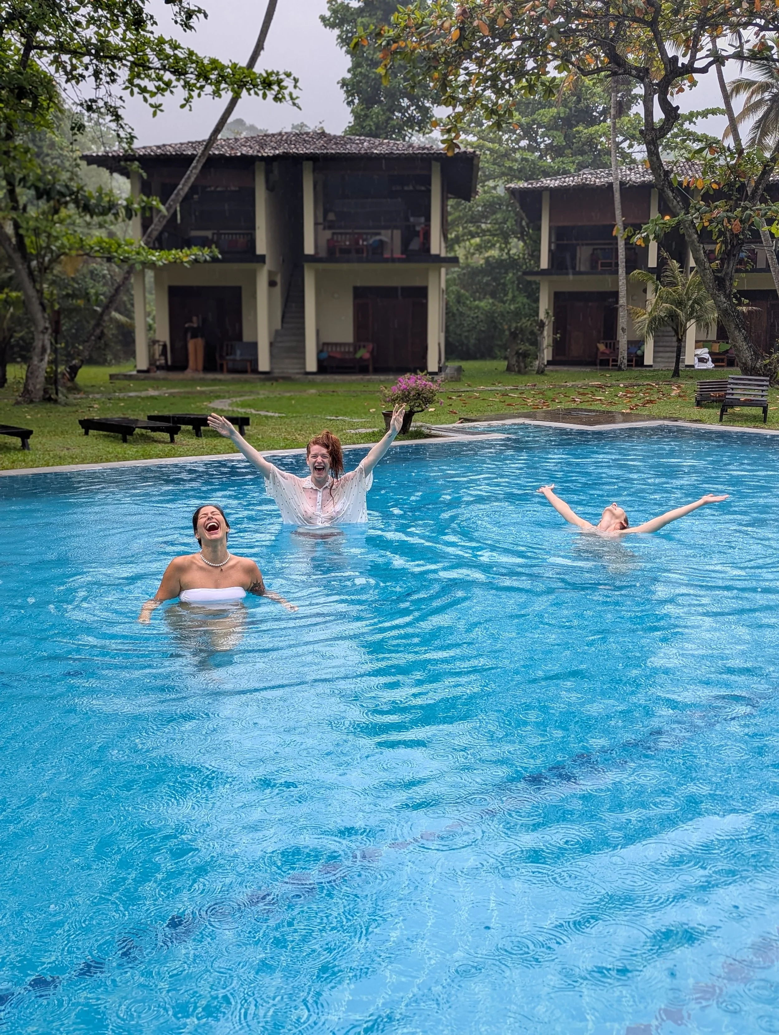 Three women having fun in a swimming pool on a rainy day, with tropical trees and two houses in the background.