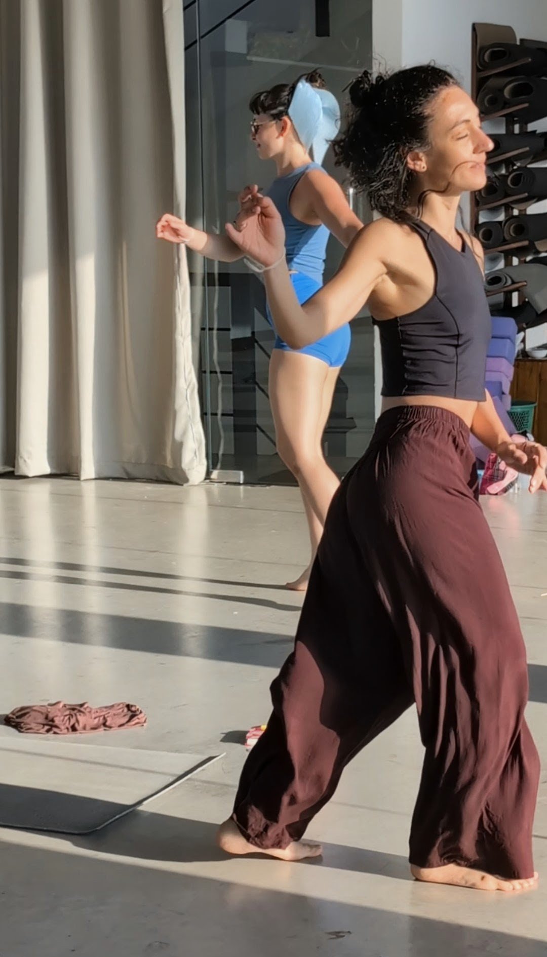 Two women are dancing barefoot in a studio, one in a black tank top and maroon loose pants, the other in a blue athletic outfit with a towel on her head, with a large window and shelves with rolled yoga mats in the background.