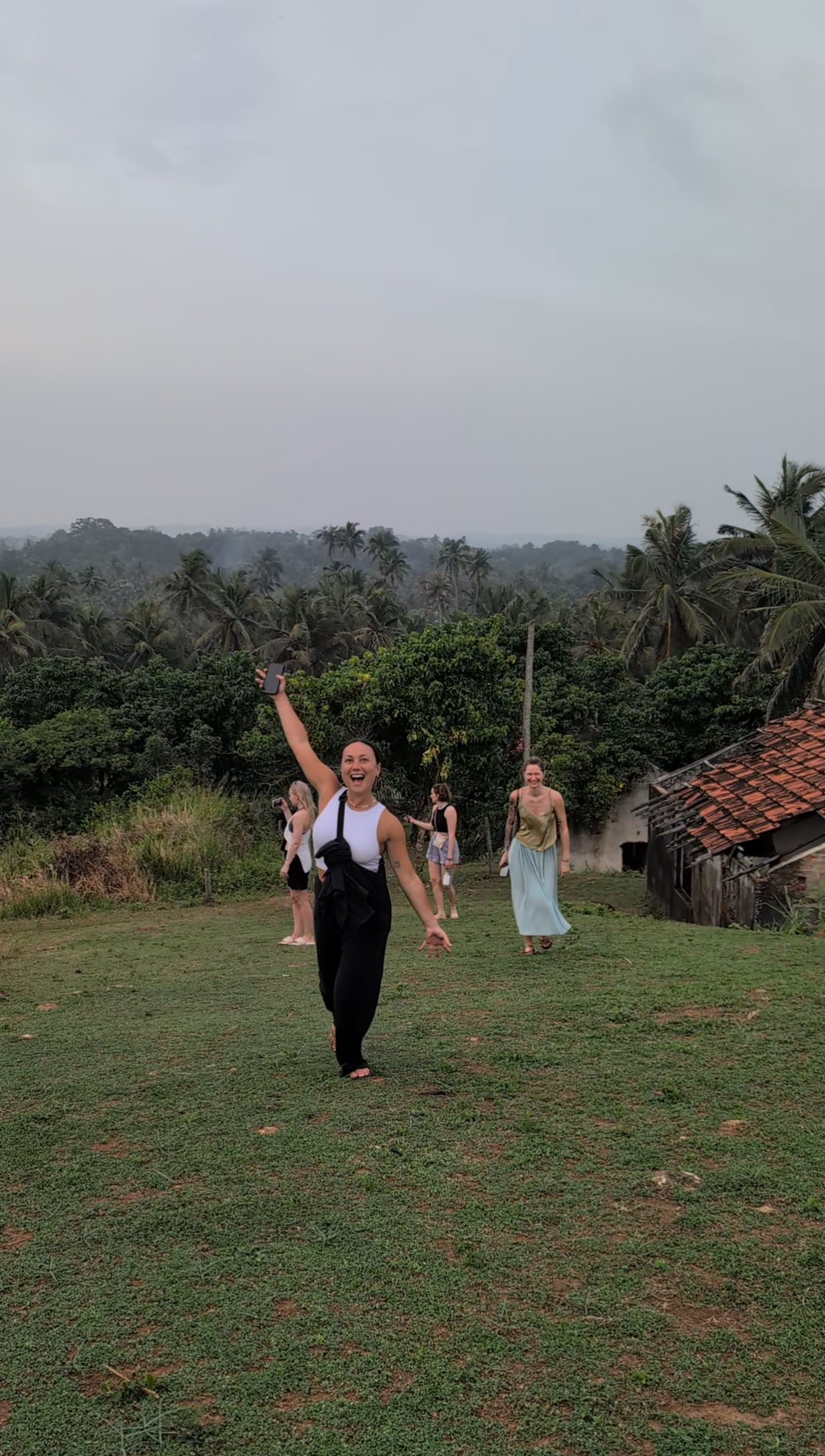 A woman smiling and holding her phone in the air, walking on a grassy area with other women in the background, surrounded by lush greenery and trees, under a cloudy sky.