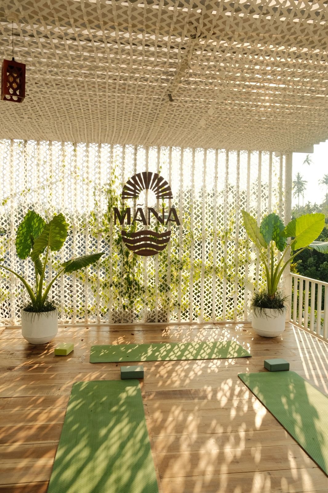 Yoga mats and blocks on a wooden floor in a sunlit outdoor space with potted plants and a decorative white lattice wall, featuring a sign that says 'MANA' with a sun and wave design.