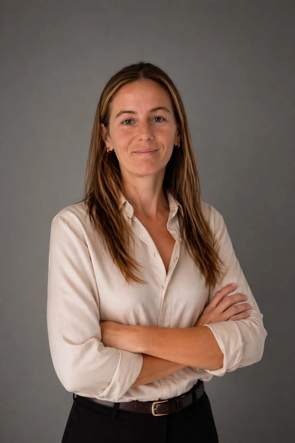 Retrato de una mujer con camisa de seda color crema, cabello largo y castaño, con brazos cruzados, sonriendo ligeramente, contra un fondo gris.